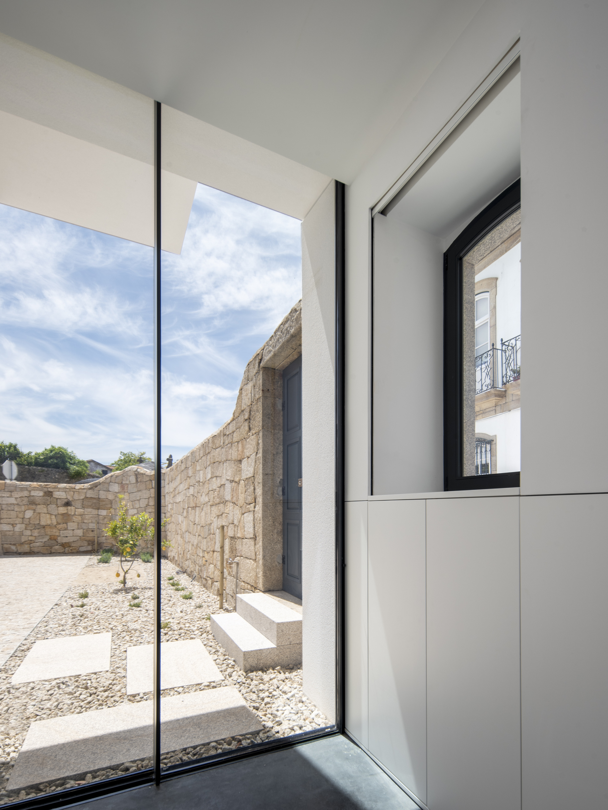 Floor-to-ceiling glass corner looking onto a granite wall, stone steps and a gravel courtyard with stepping slabs and a small tree under scattered clouds