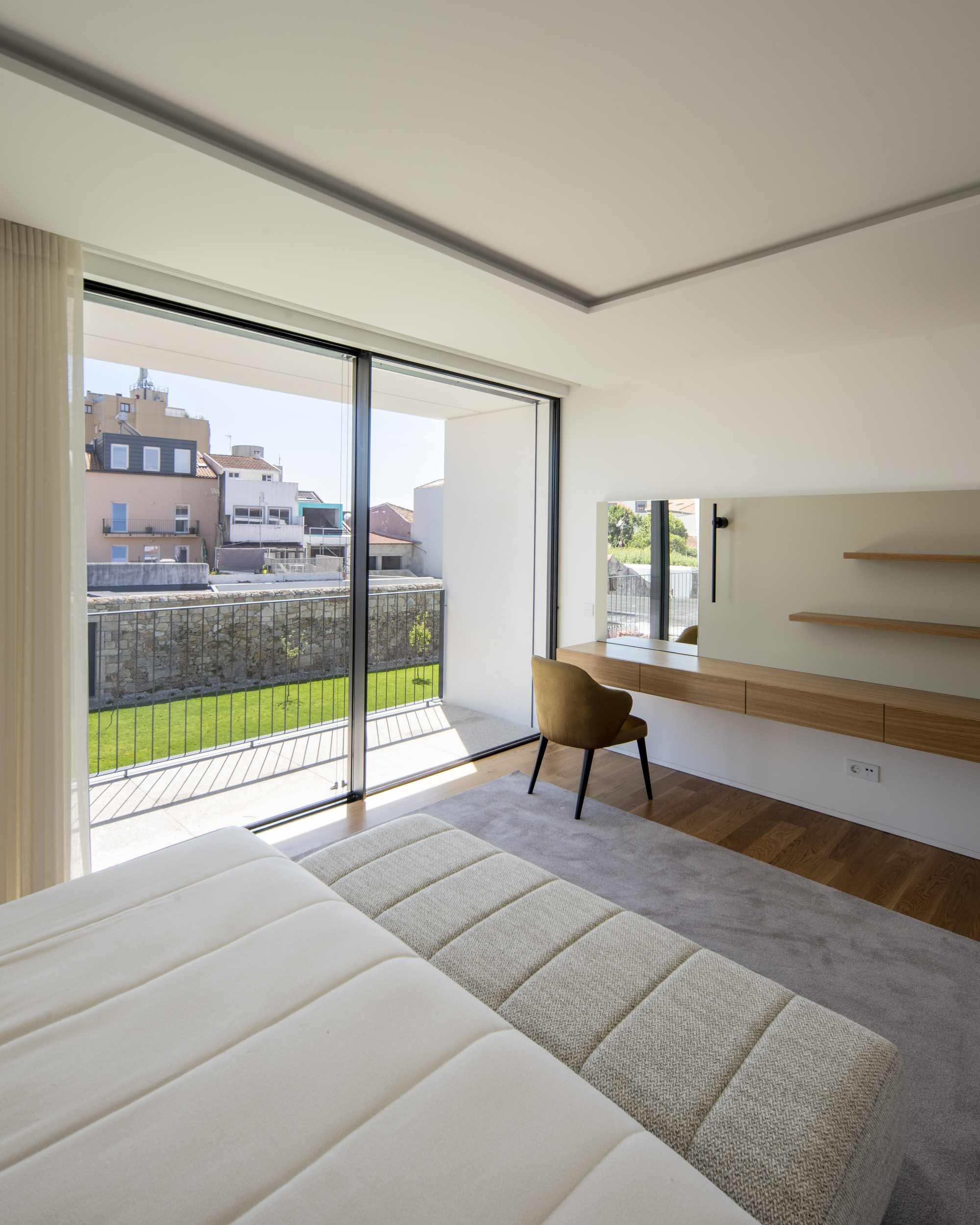 Bedroom with an upholstered bench, timber dressing shelf and ochre chair, opening through sliding glass doors to a balcony above a lawn in daylight
