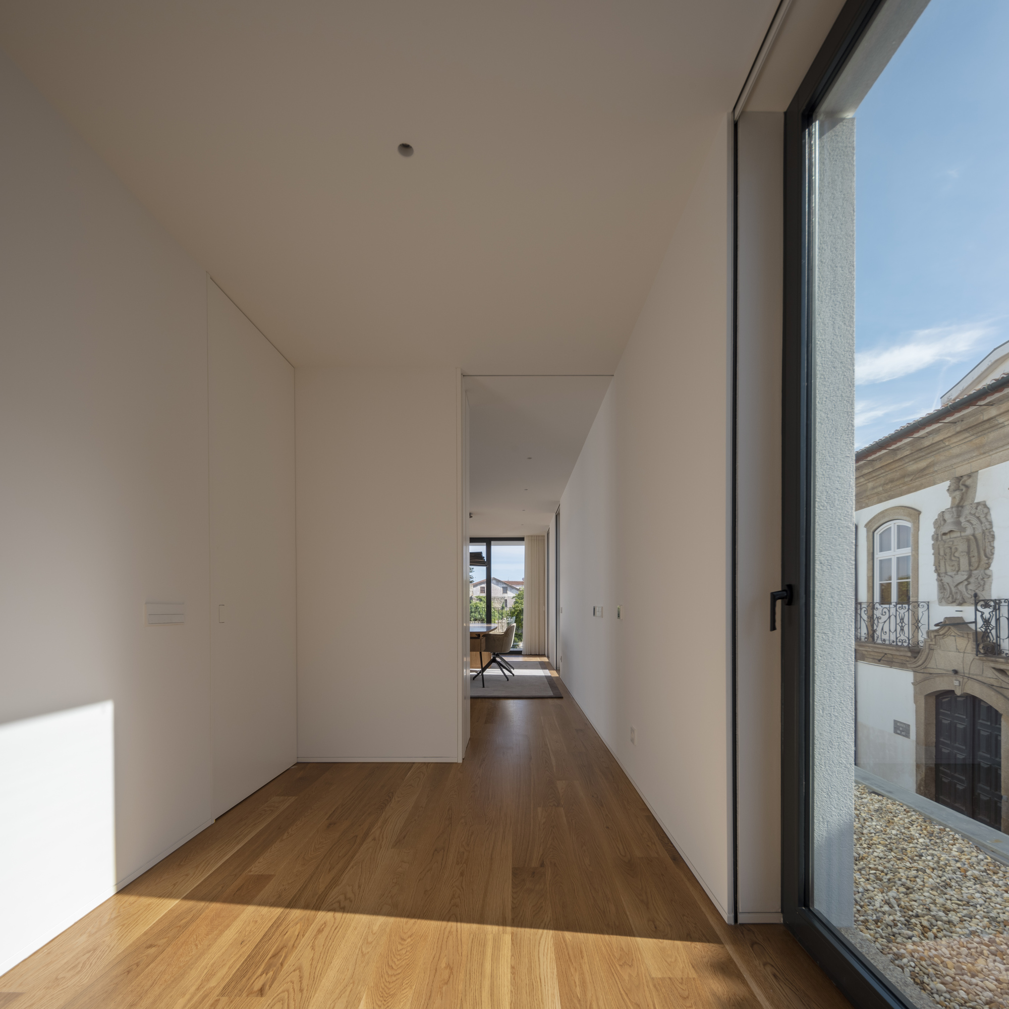 Narrow white corridor with oak floor and full-height window overlooking a gravel court and an ornate stone facade in afternoon sun