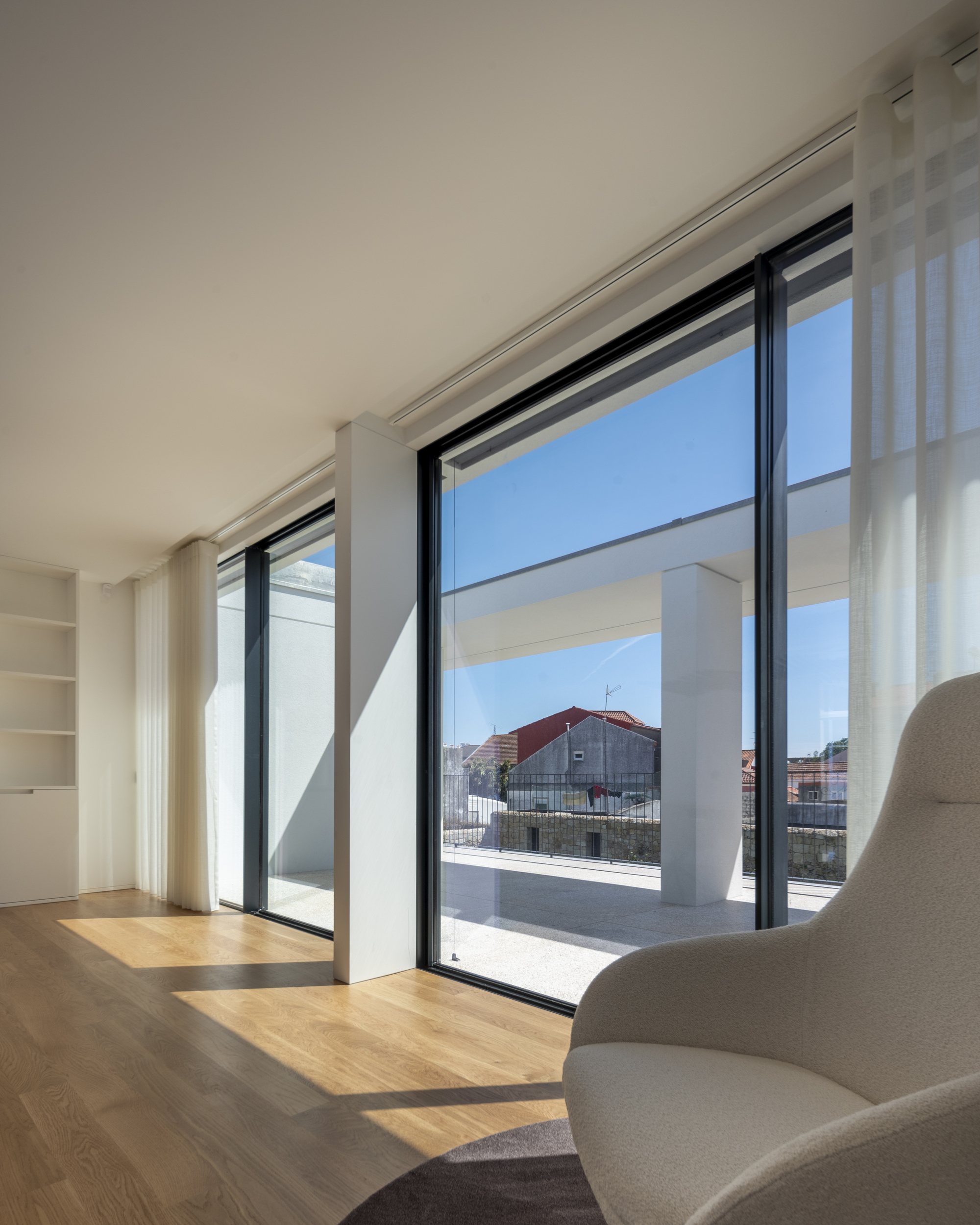 Floor-to-ceiling black-framed glazing with sheer curtains and a boucle armchair, looking onto a terrace column and neighboring red roofs under blue sky