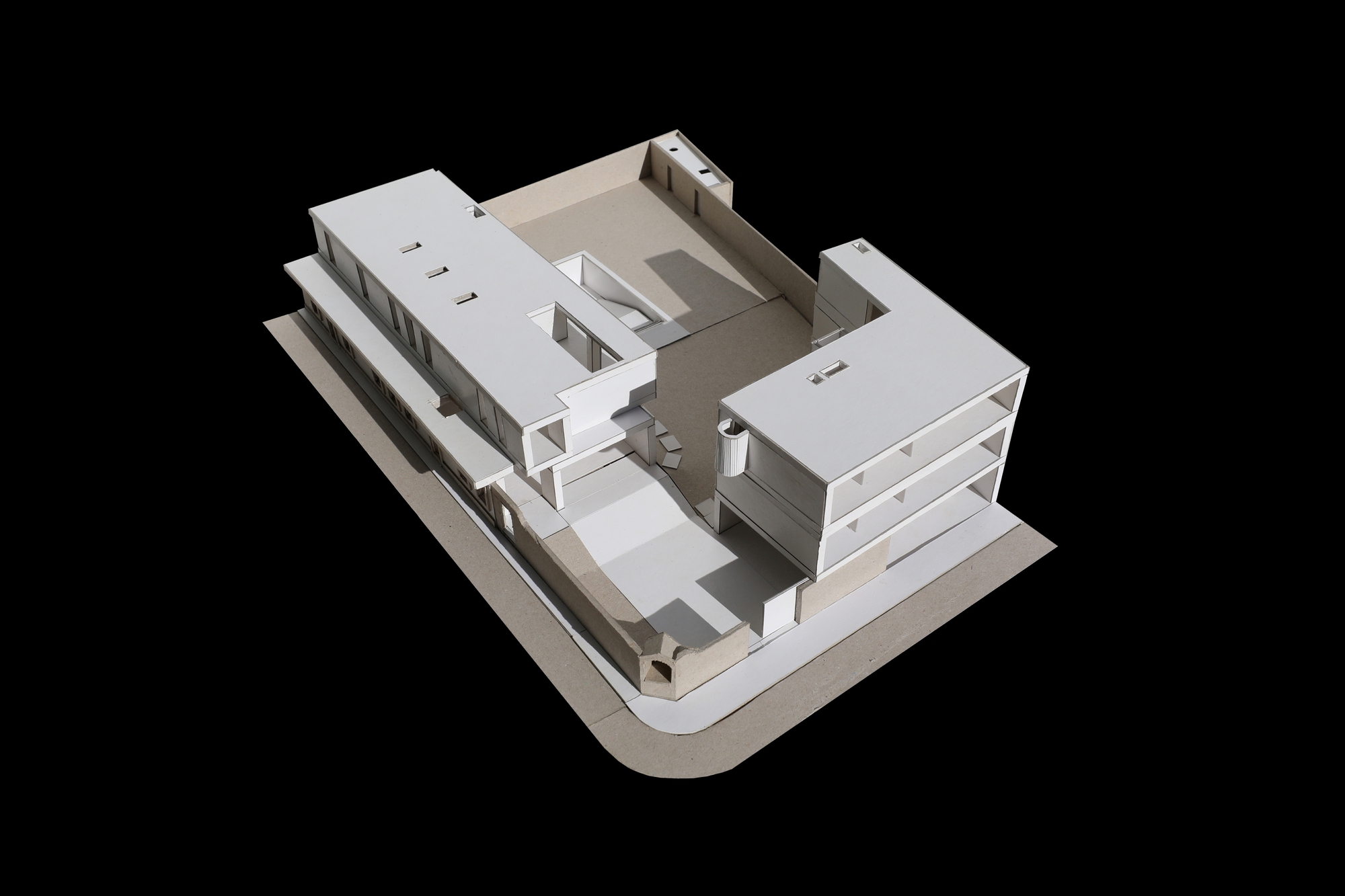 Aerial view of a white card model showing an L-shaped arrangement of stacked volumes around a sunken courtyard on a corner plot