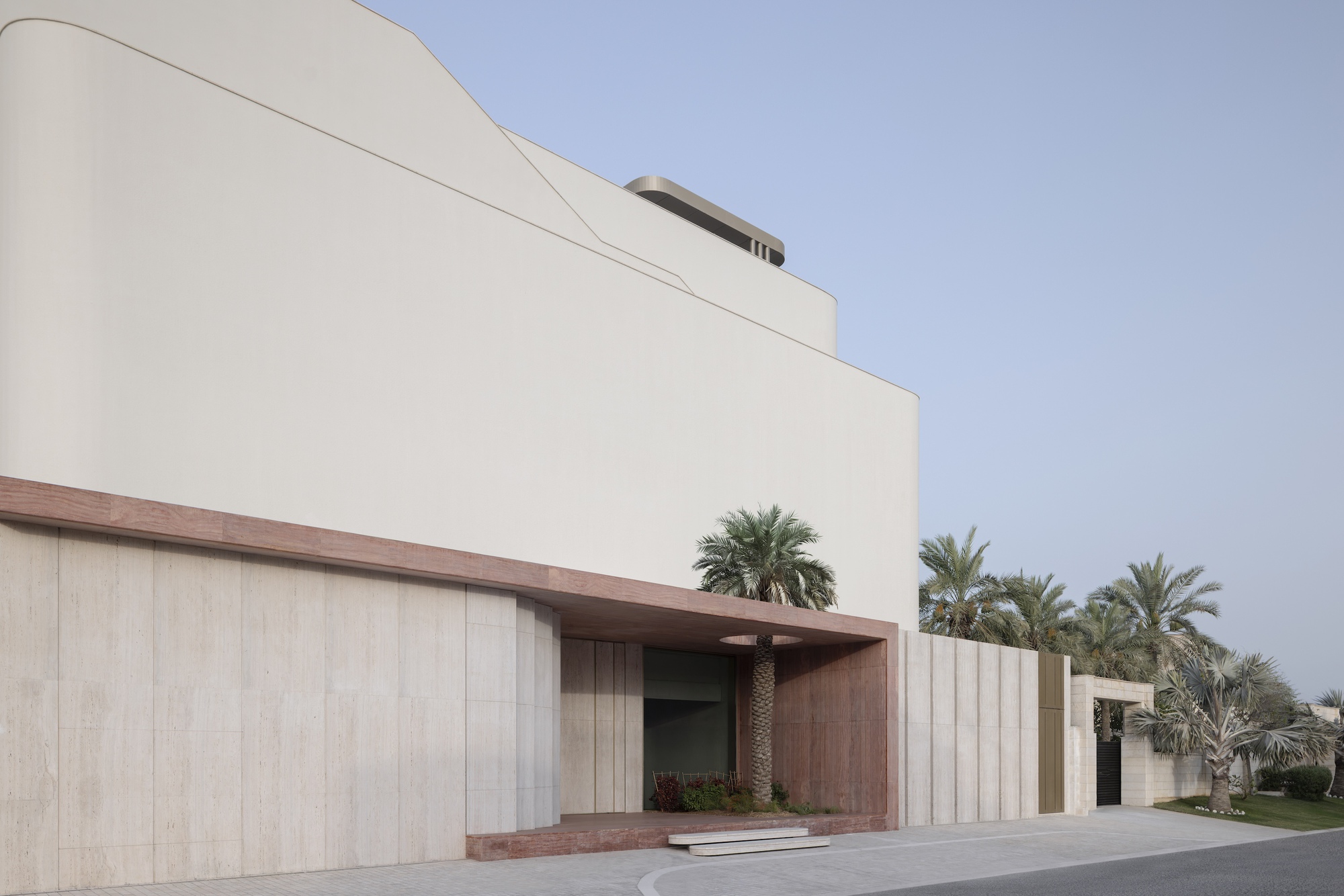 Street facade with travertine panel walls and a pink stone canopy sheltering a palm at the entrance, under a clear dusk sky