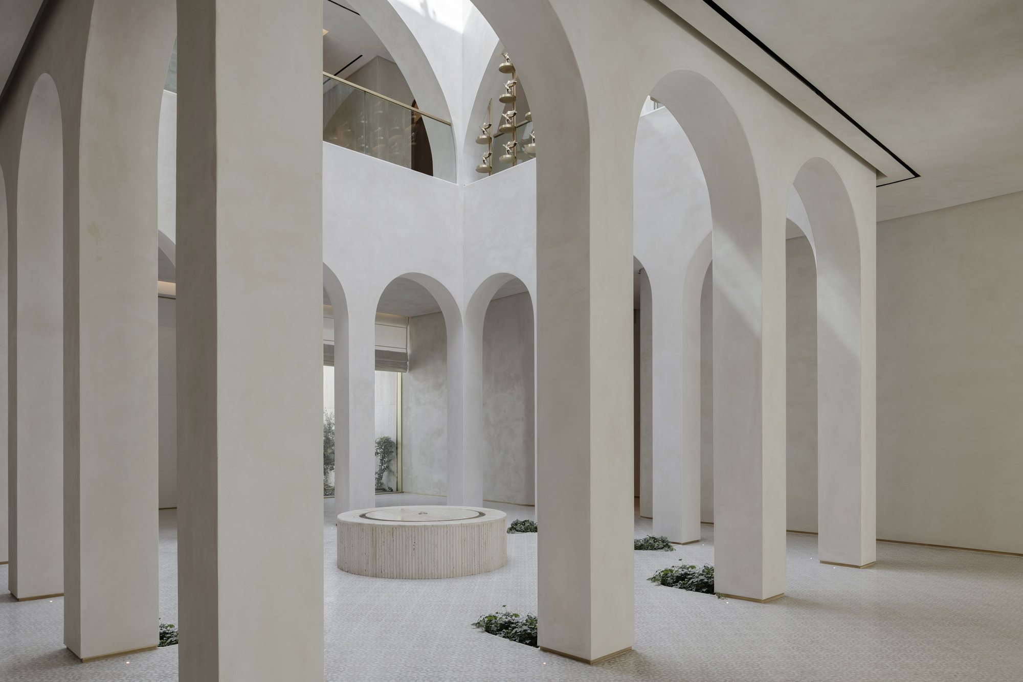 Arched columns around a round stone fountain in a pale plaster atrium, with soft daylight falling across the floor