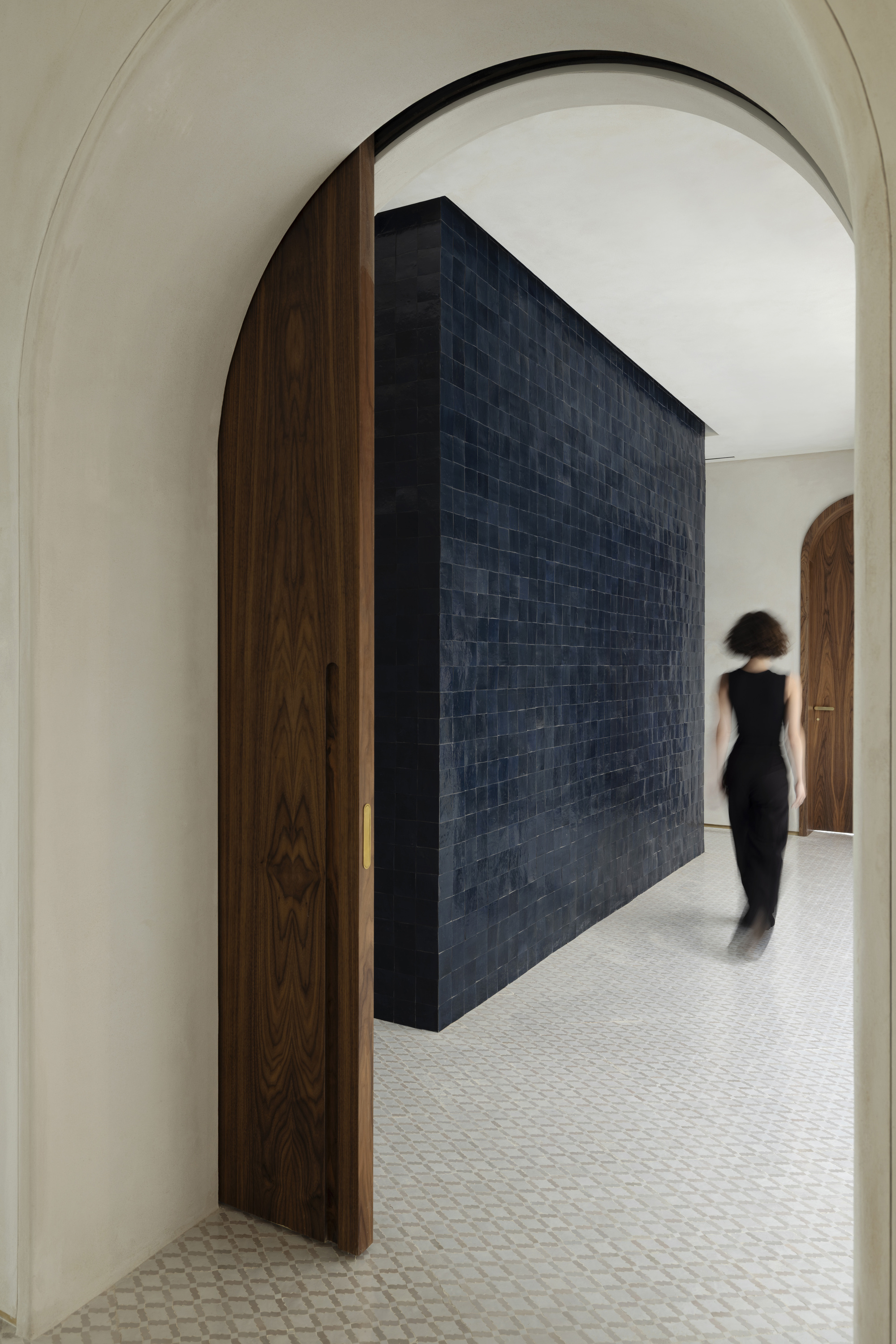 Walnut arched door opening onto a dark blue glazed tile volume as a blurred figure walks past