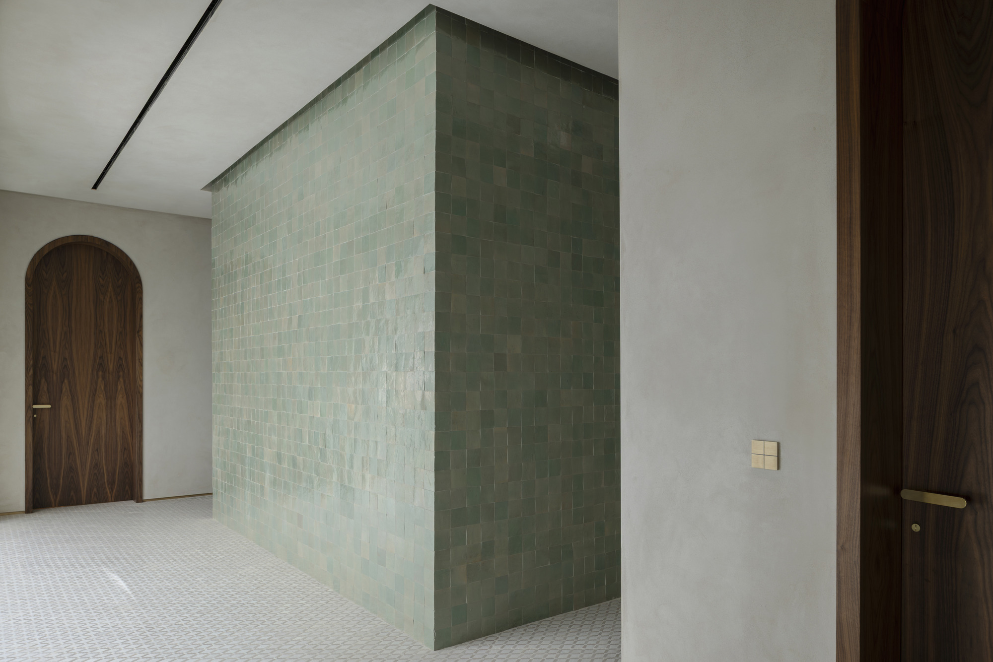 Corner of a green glazed tile volume beside a walnut arched door and a brass switch plate