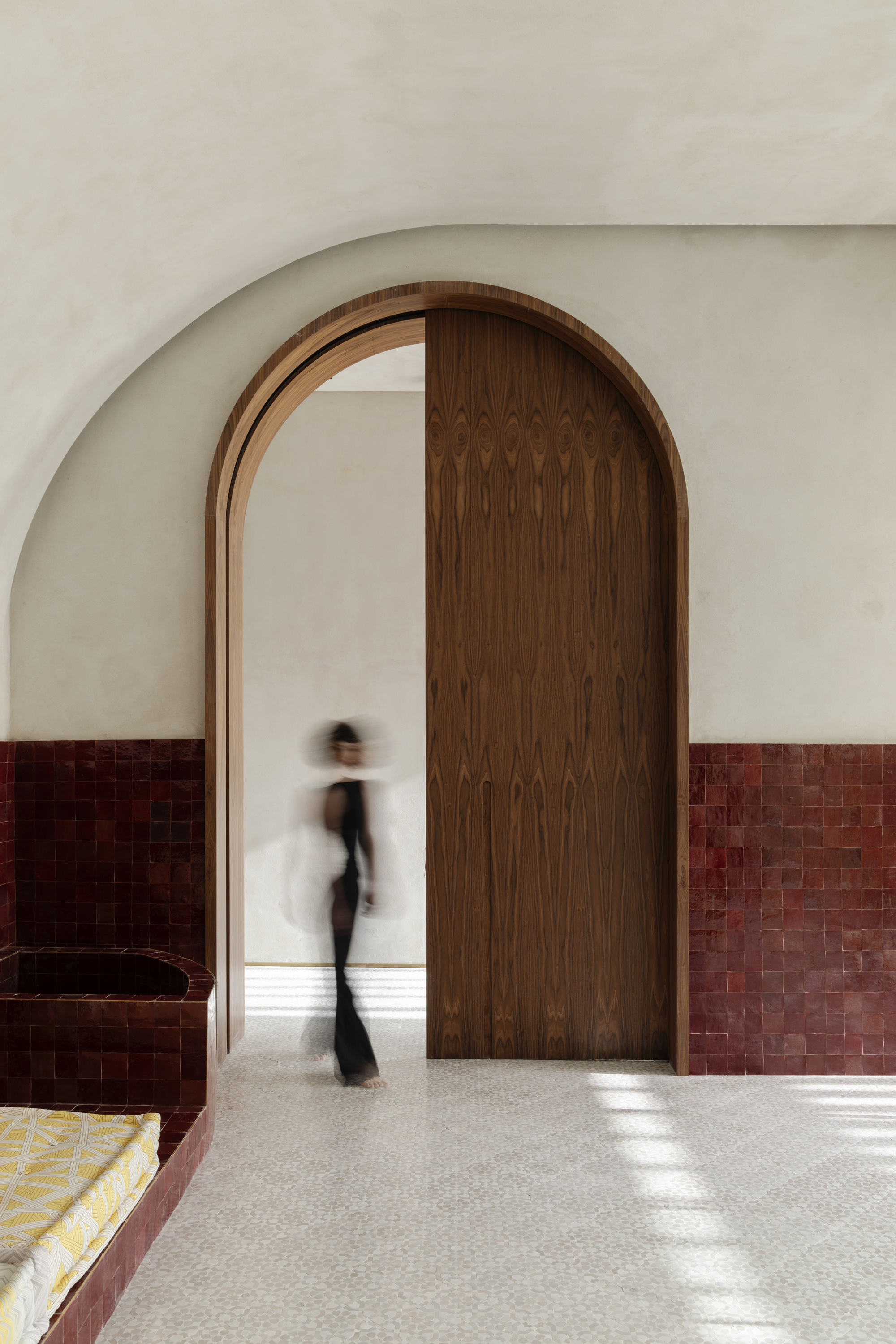 Tall walnut arched doorway between deep red tile walls, with a blurred figure passing and sunlight patches on the floor