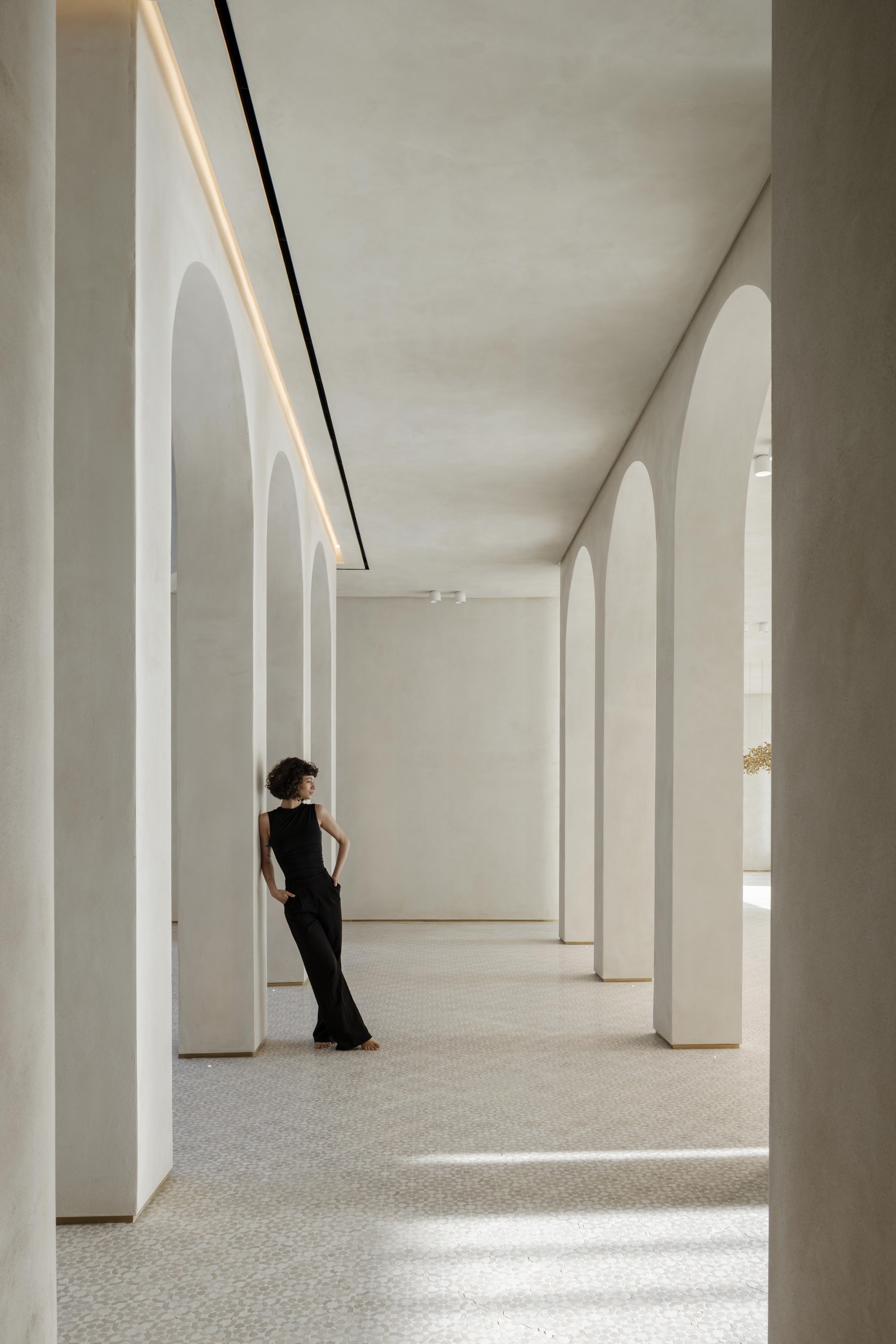 Colonnade of plaster arches with a linear light strip, a woman in black leaning on a column in raking light