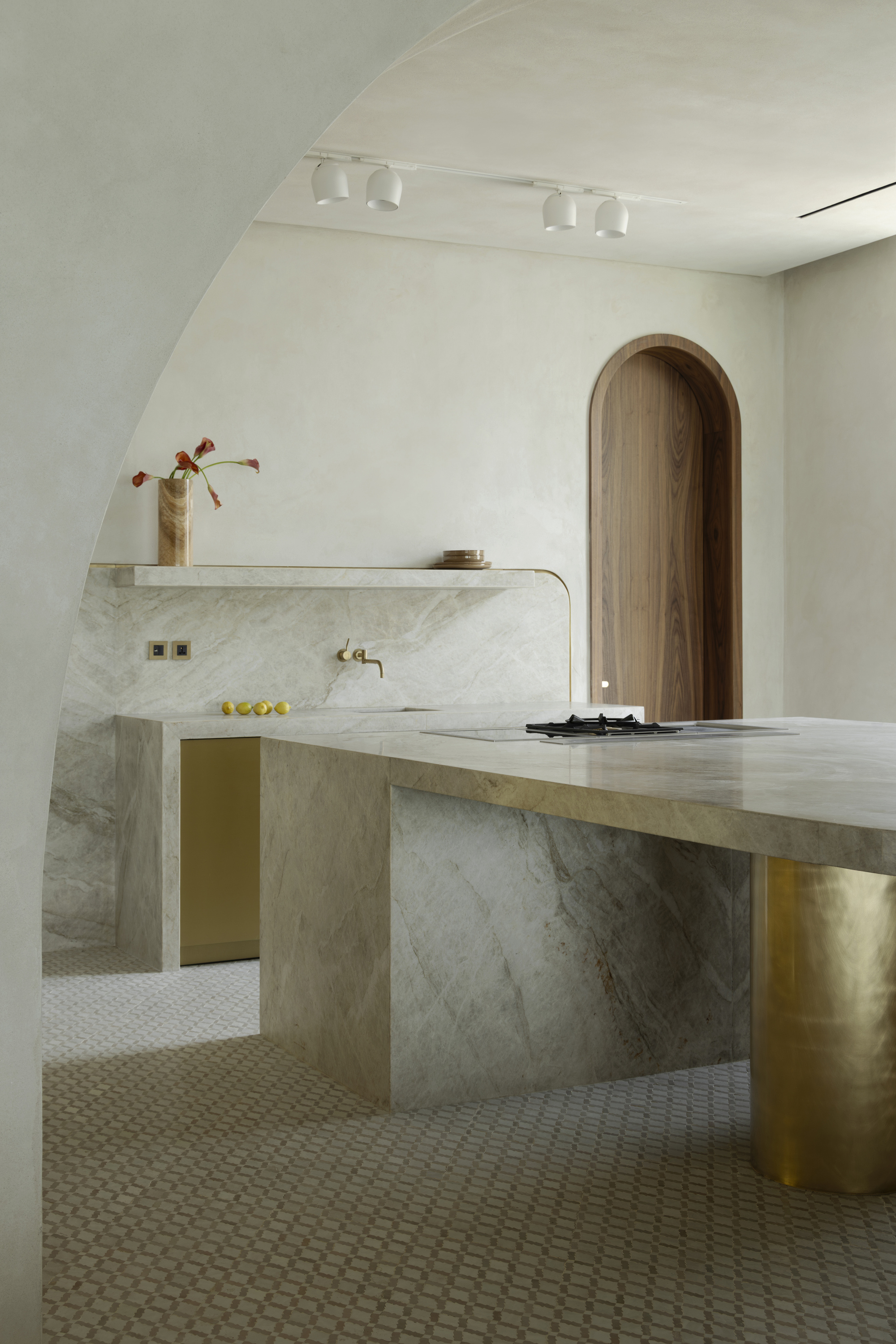 Stone kitchen island with brass cylinder leg beside a marble backsplash and an arched walnut door, lit by ceiling spotlights