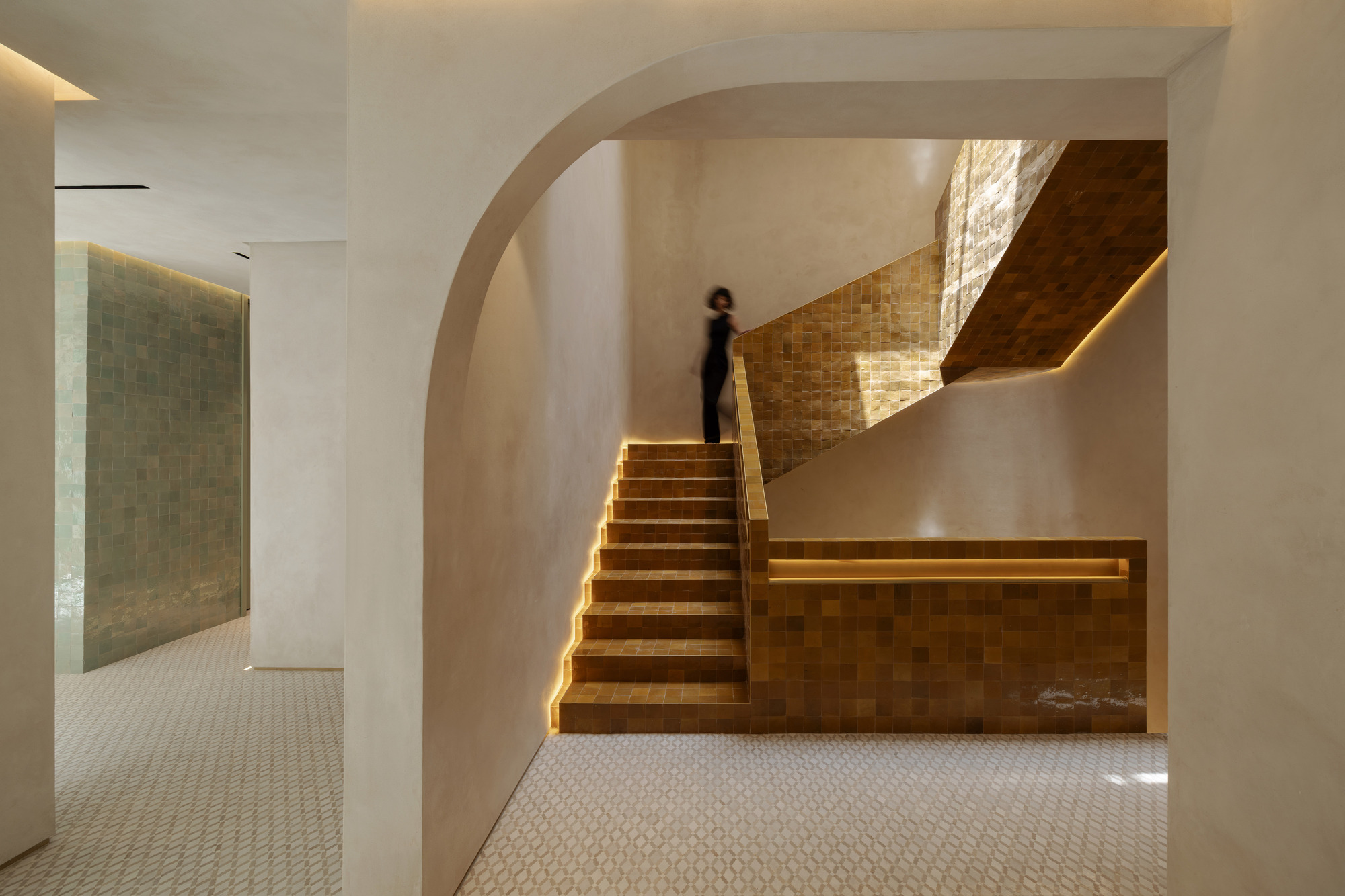 Staircase clad in amber glazed tiles beneath a plaster arch, with a blurred figure climbing in warm edge light