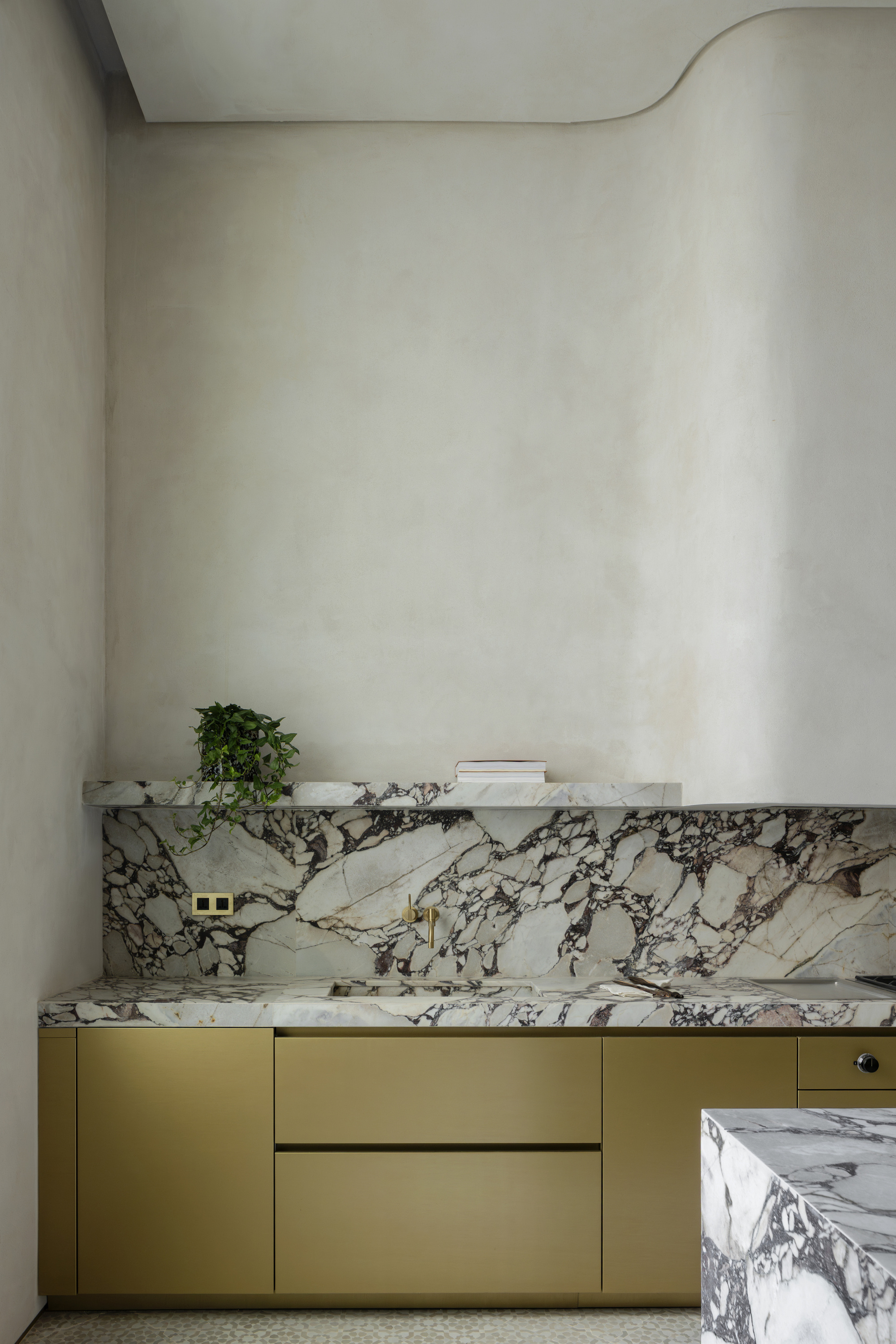 Kitchen counter with bold veined marble backsplash and brass cabinet fronts under a curved plaster wall, a trailing plant on the shelf