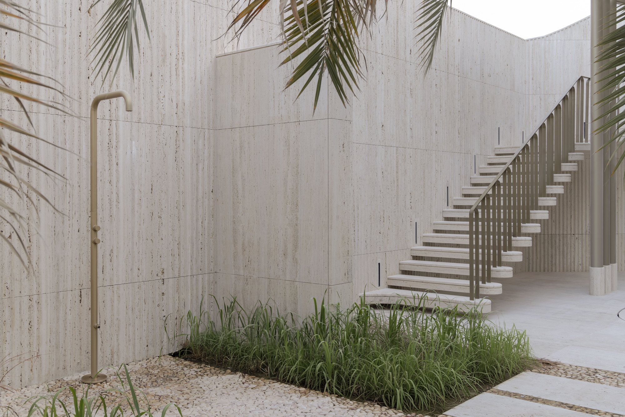 Travertine-clad courtyard with a brass outdoor shower, a floating stair with slim balustrade and a grass bed under palm fronds