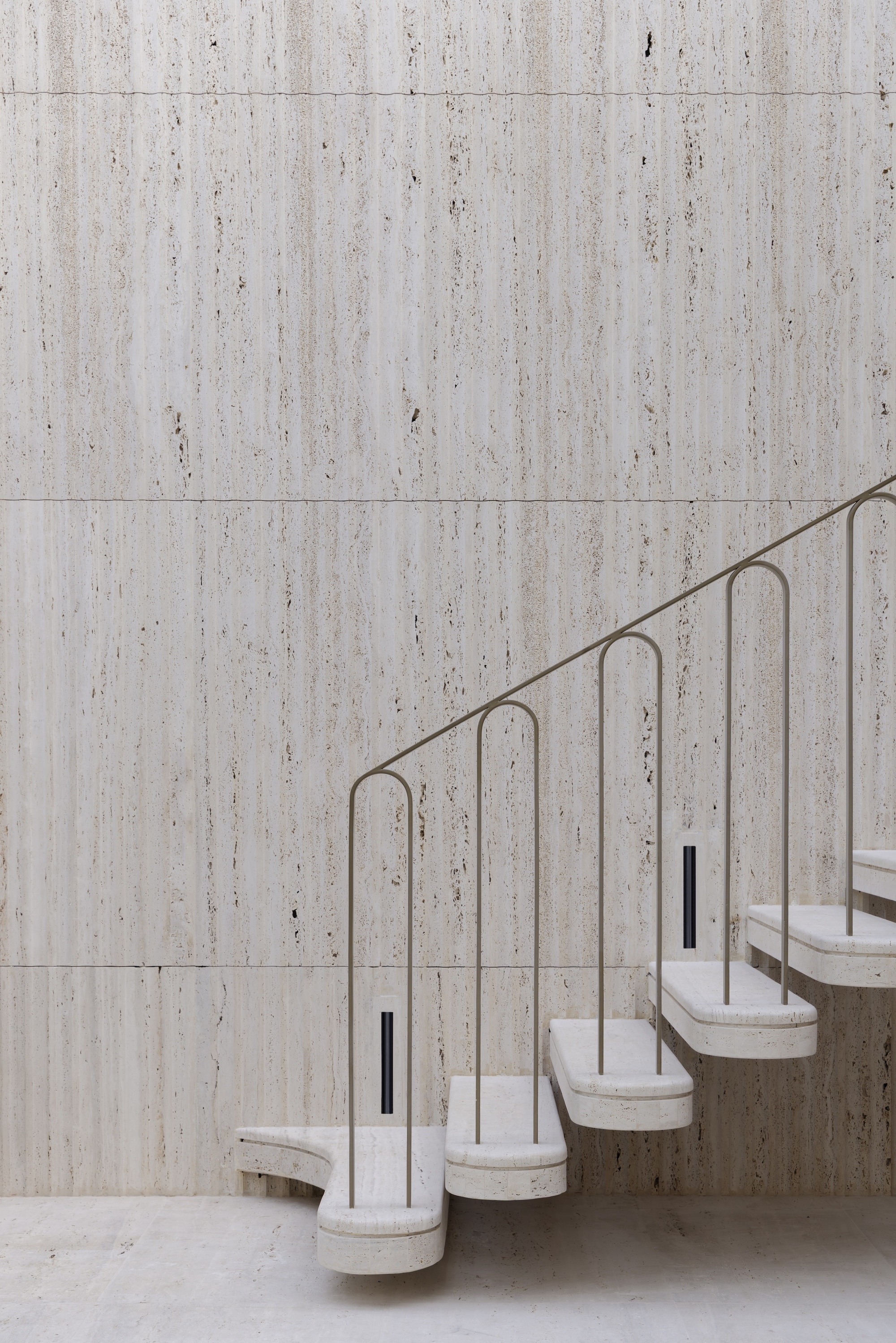 Cantilevered travertine stair with slim looped metal balustrade rising against a tall travertine-clad wall in soft even light