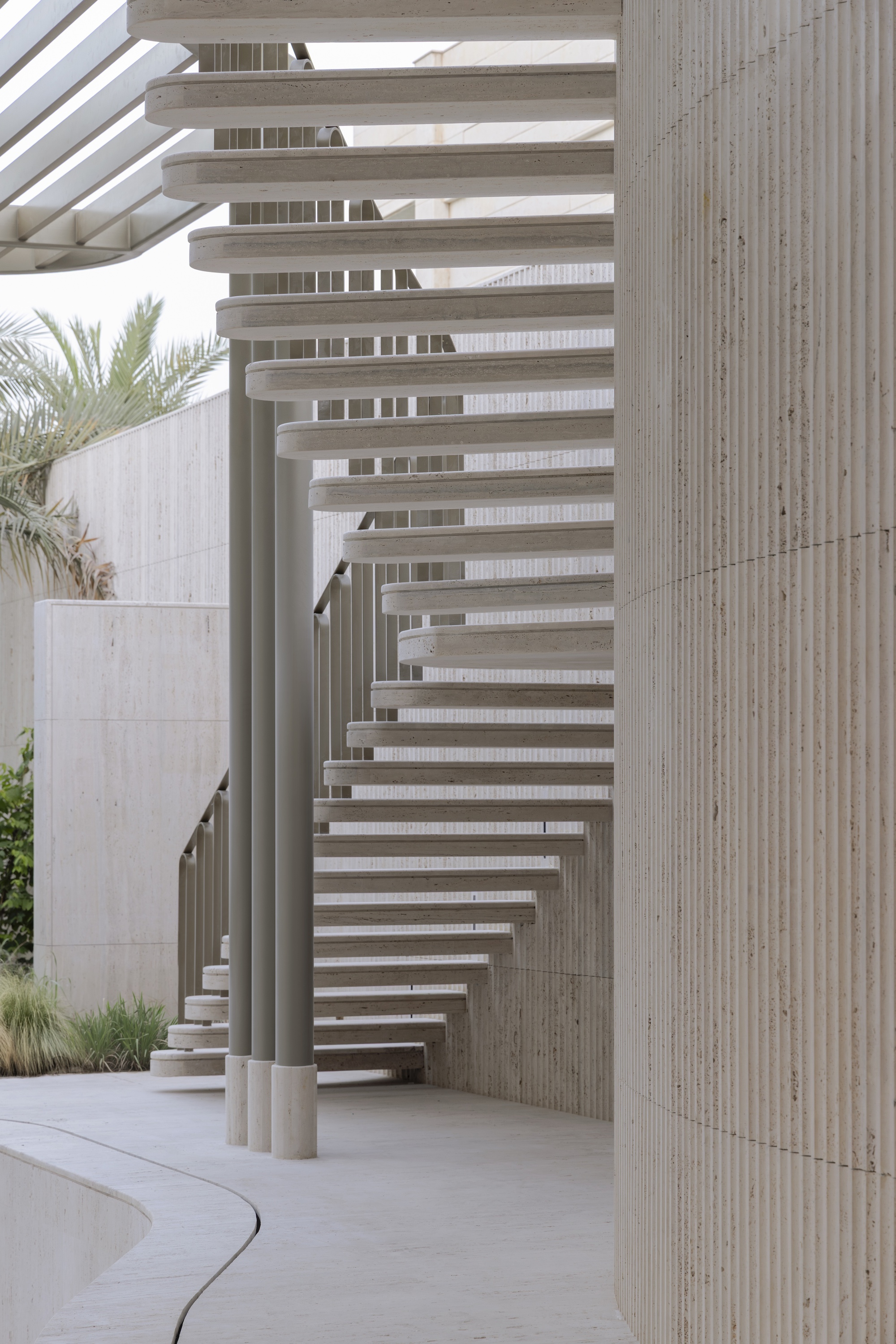 Floating stair treads beside a fluted stone curved wall, with palm fronds and a slatted pergola overhead in daylight