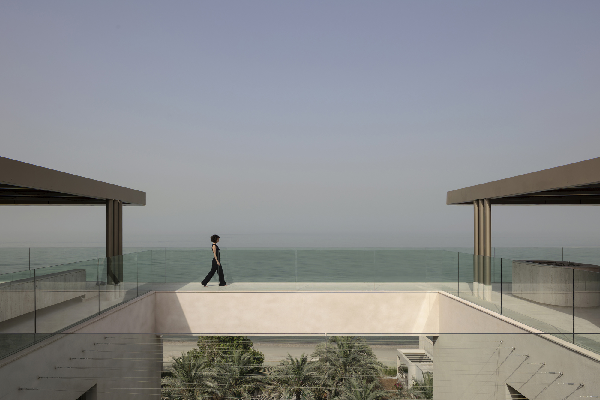 Woman walking across a roof terrace edged with glass balustrades, with the sea and palm tops beyond in hazy light