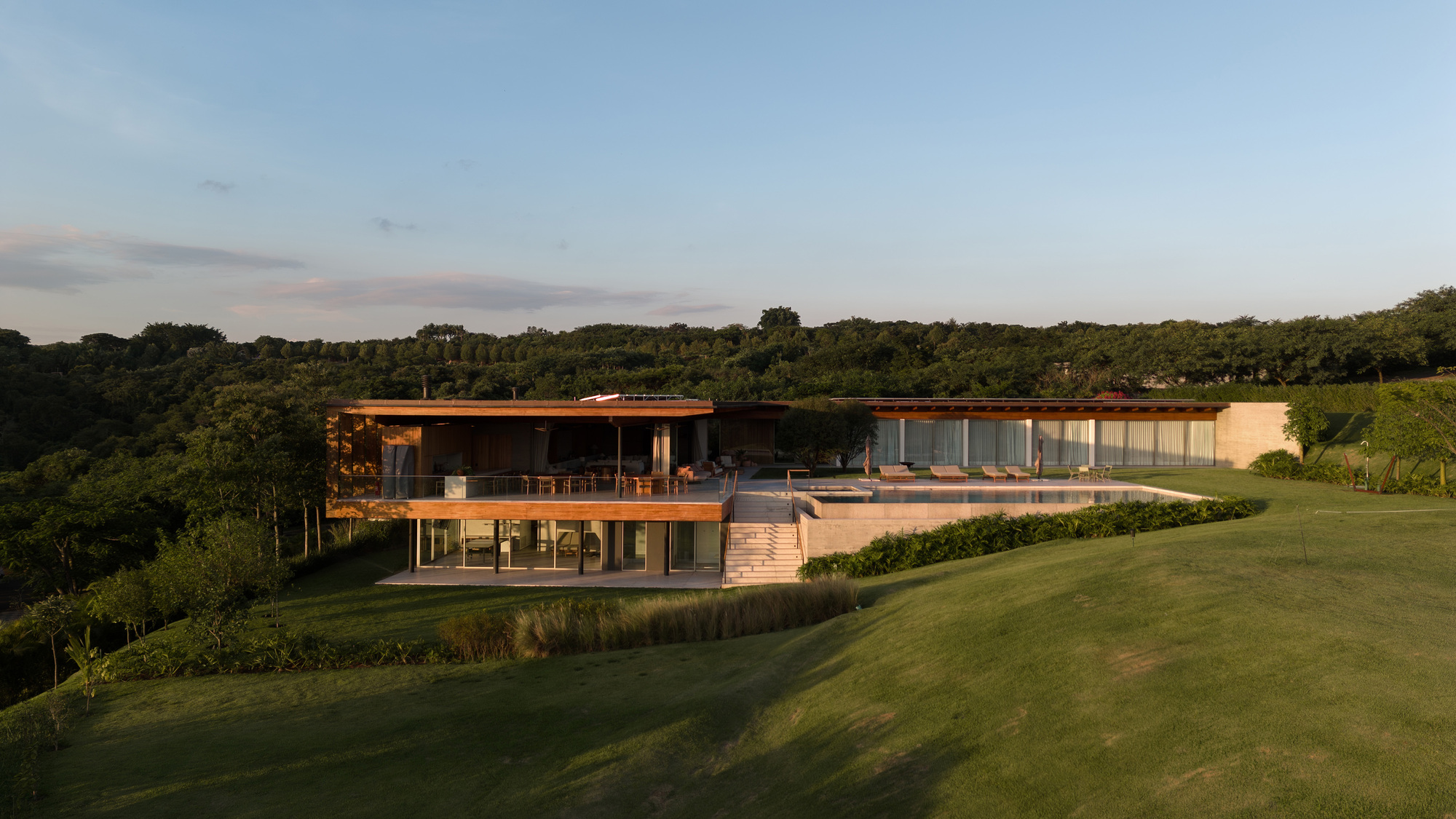 Long timber-roofed house with a raised terrace, stair and infinity pool above a grassy slope in low evening light