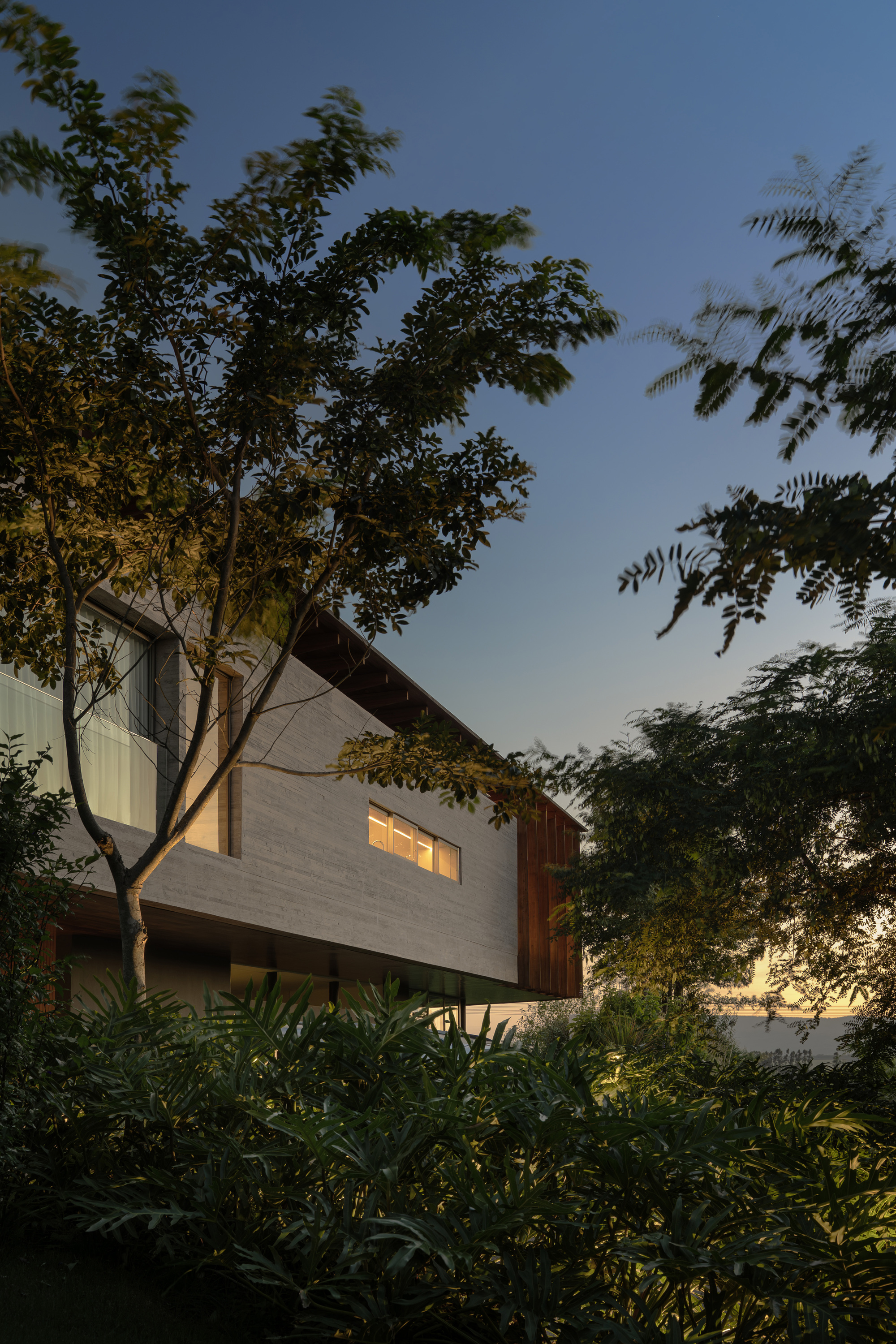 Upper volume of banded concrete and rust-toned cladding with a lit strip window, seen through tree branches at dusk