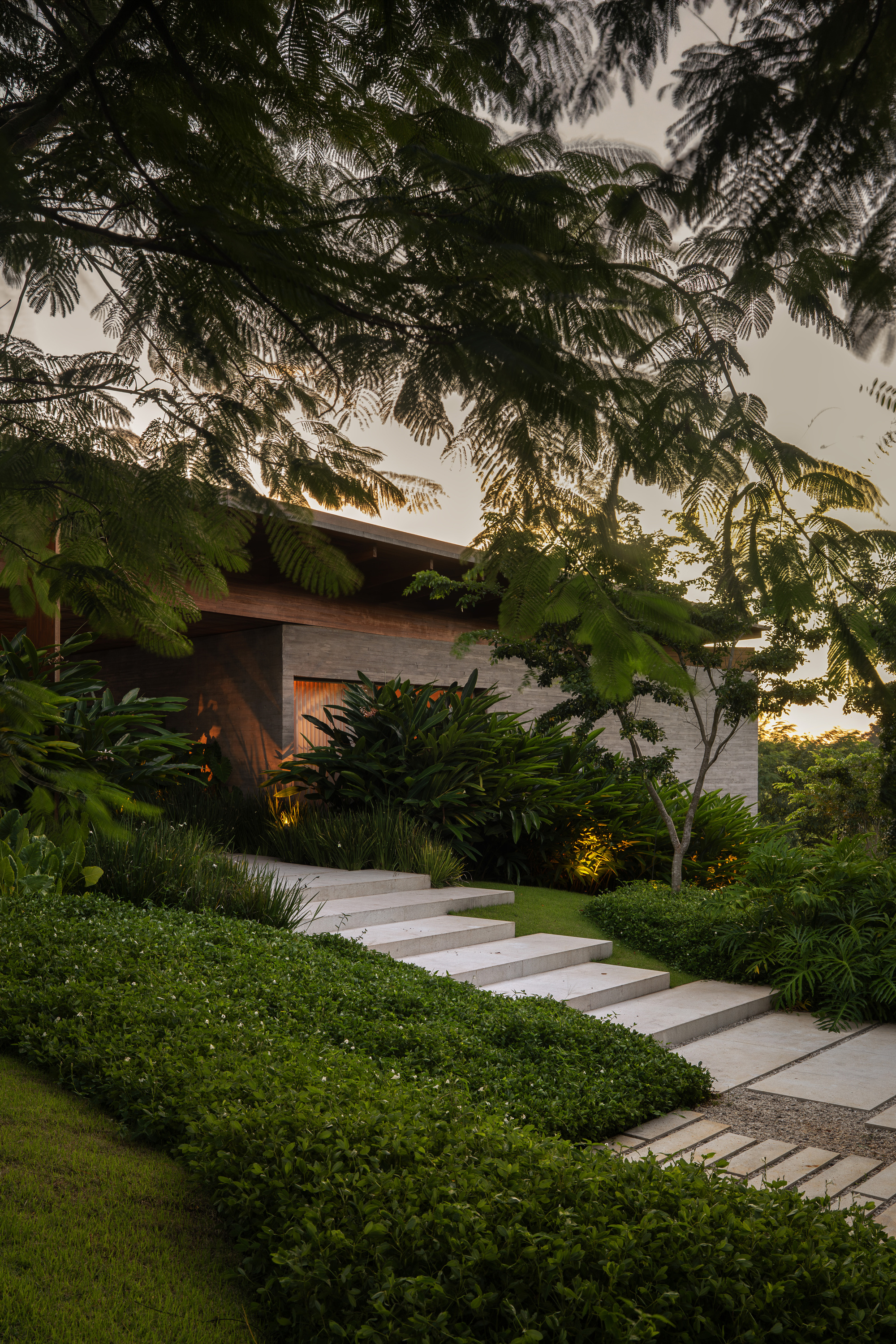 Pale stone steps rising through groundcover toward a concrete and timber pavilion, with uplights glowing among tropical plants at dusk