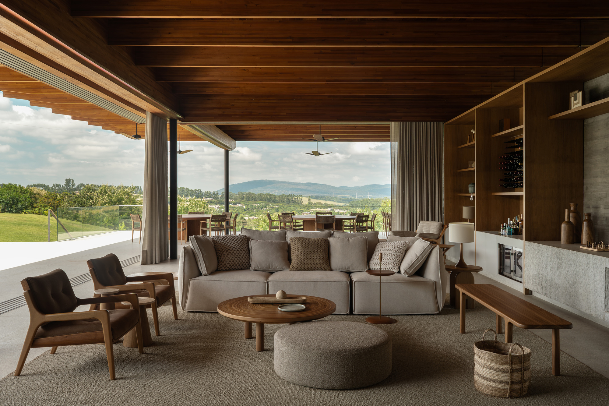 Living room with a low sofa, timber armchairs and wood shelving opening to a distant mountain view under cloud
