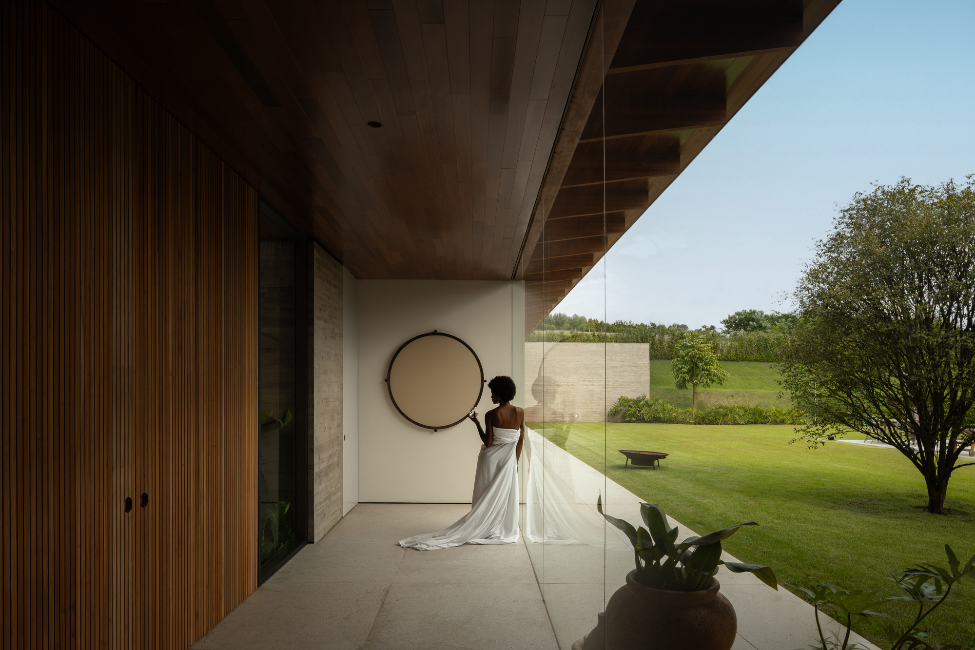 Woman in a white gown stands beside a round mirror on a covered terrace, with a glass wall facing a lawn