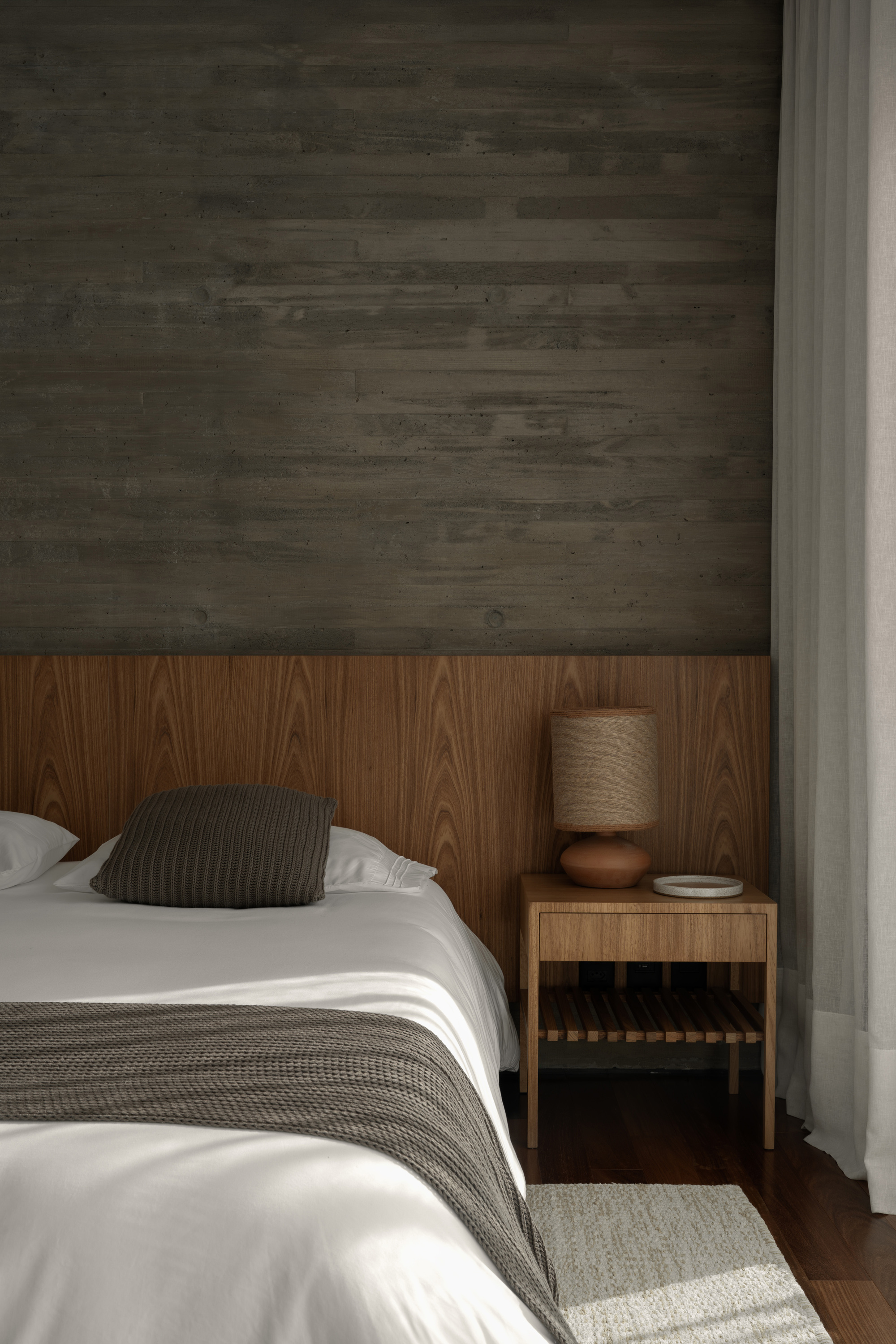 Bedroom corner with board-formed concrete wall, oak headboard panel and slatted bedside table beside a sheer curtain in soft daylight