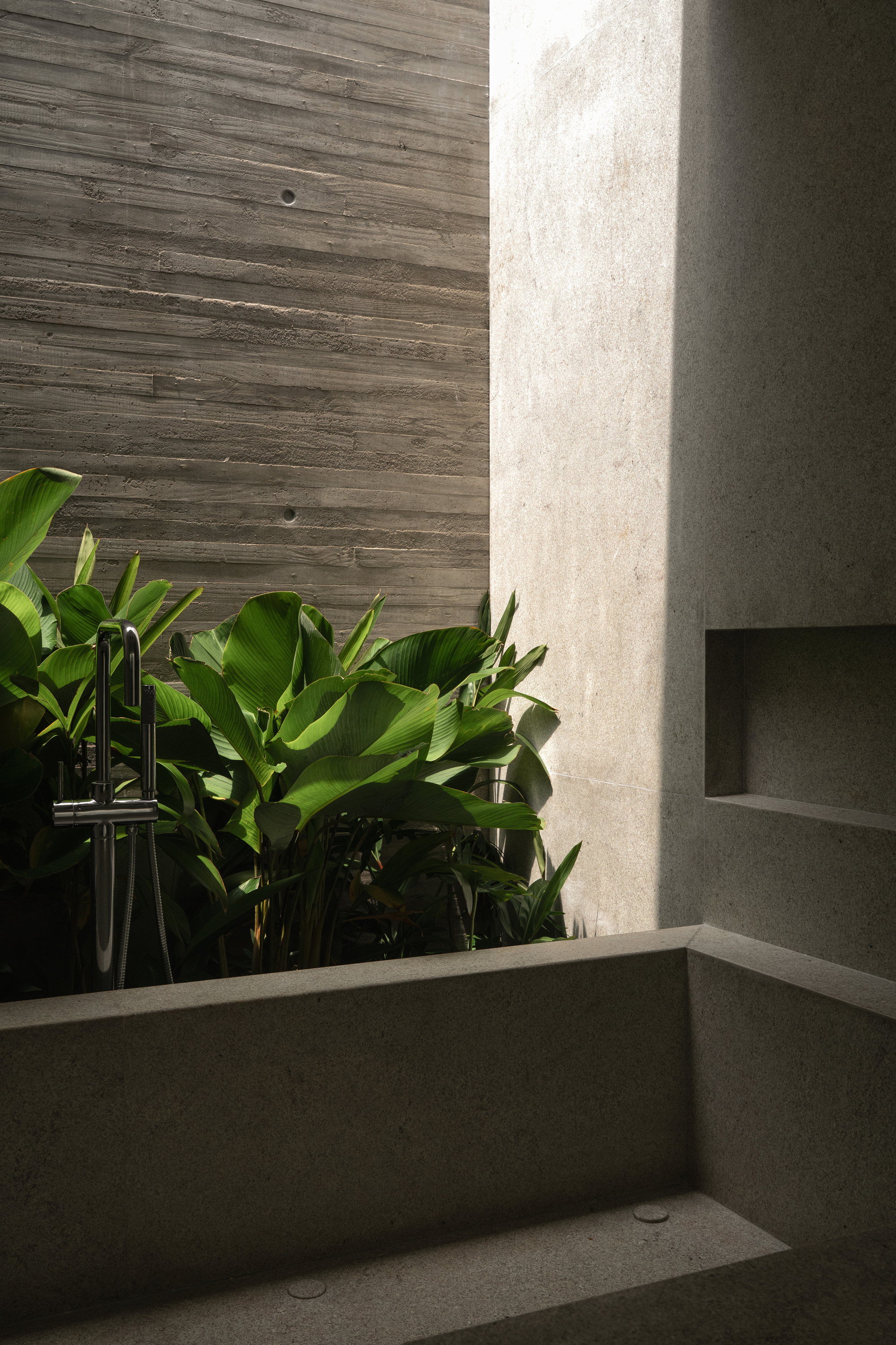 Stone bathtub beside a board-formed concrete wall, with broadleaf plants and a floor-mounted tap lit by a shaft of sunlight