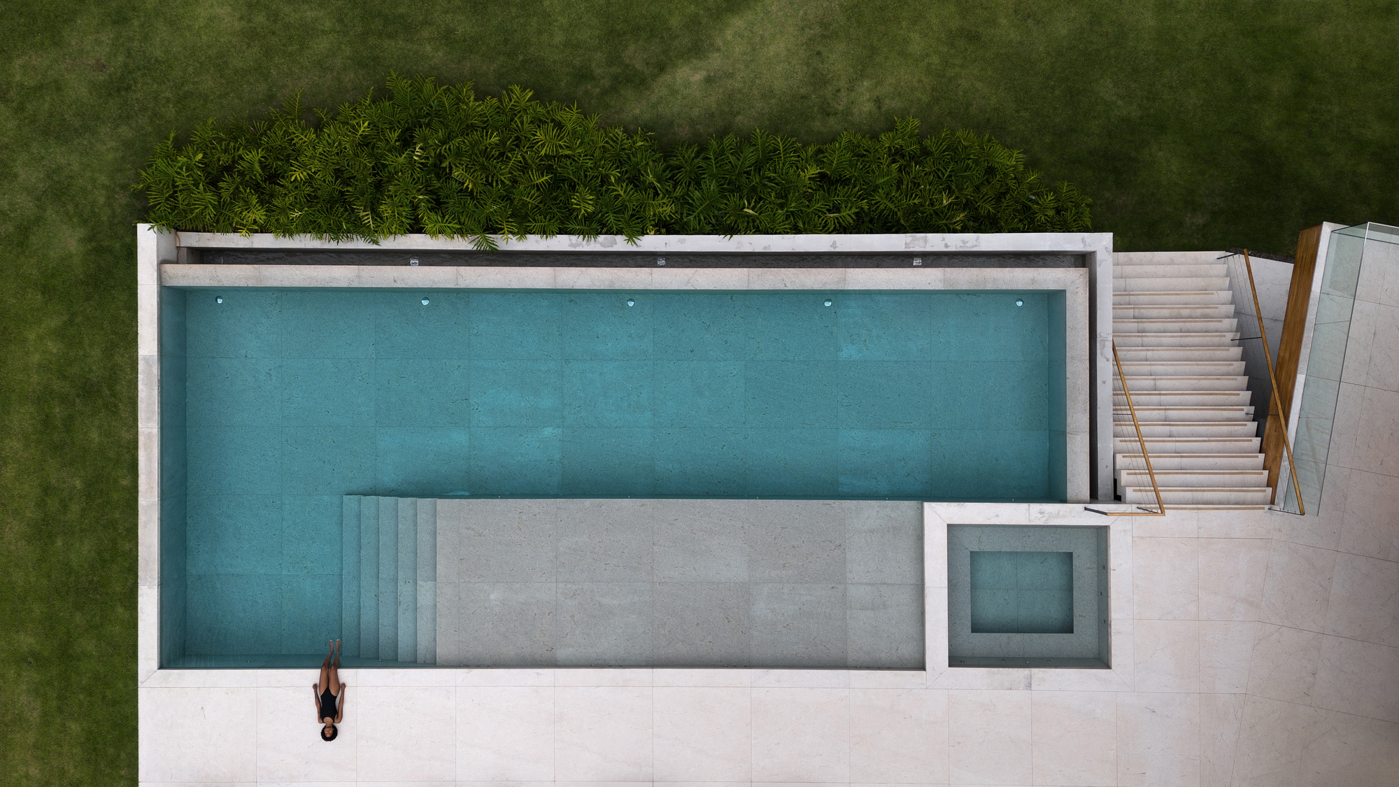 Top-down view of a rectangular turquoise pool with stone deck, steps and a small spa, with one person resting at the edge