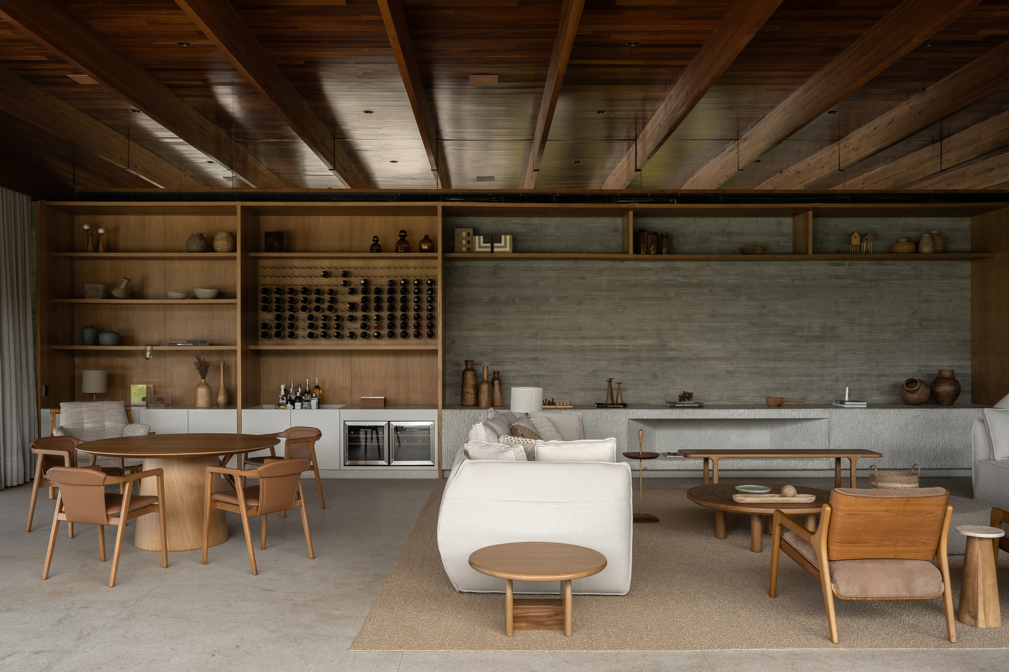 Living and dining room with exposed timber beams, built-in wine shelving, concrete wall, white sofa and round oak tables under warm light