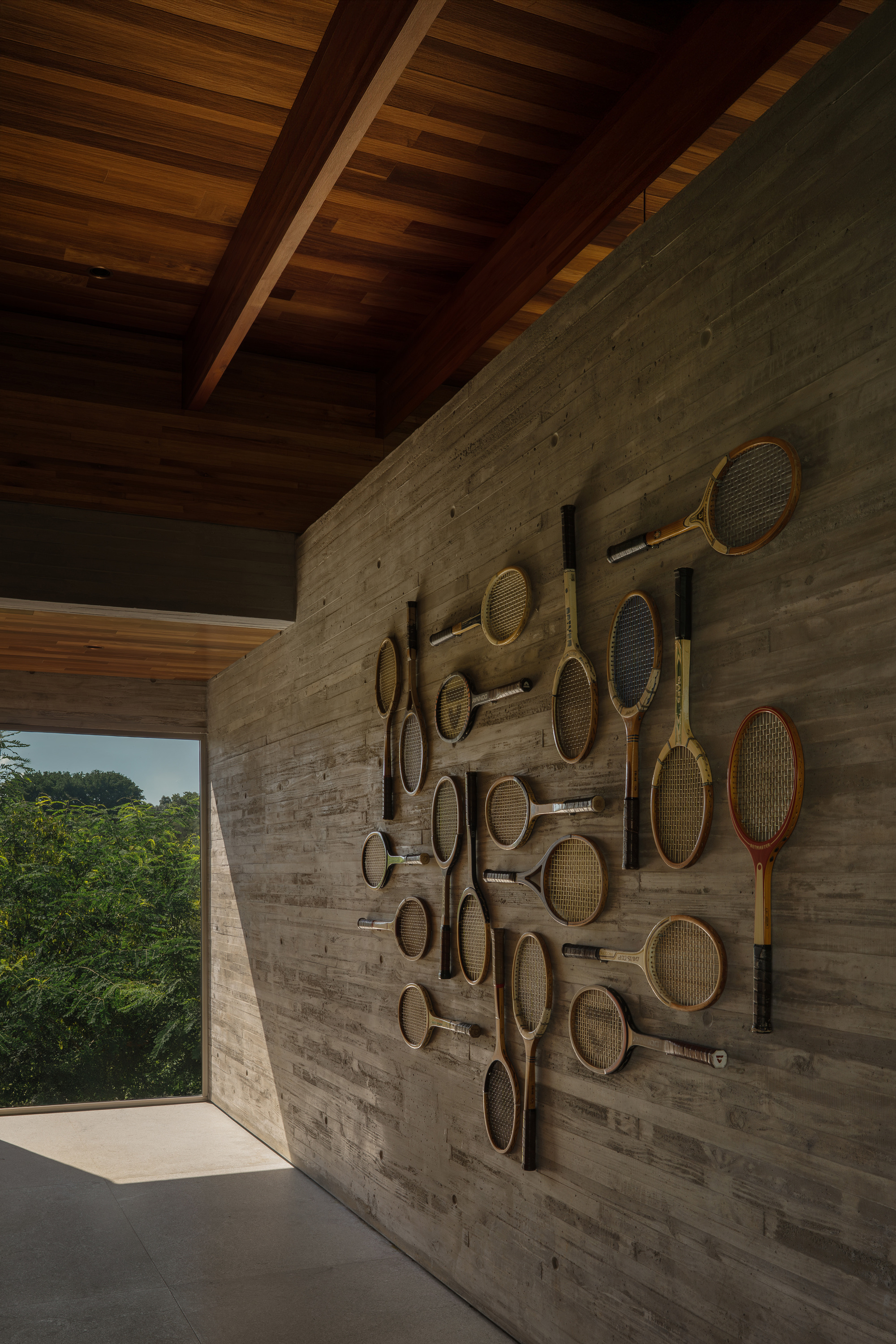 Vintage wooden tennis rackets arranged on a board-formed concrete wall beside an open corner framing green treetops in daylight