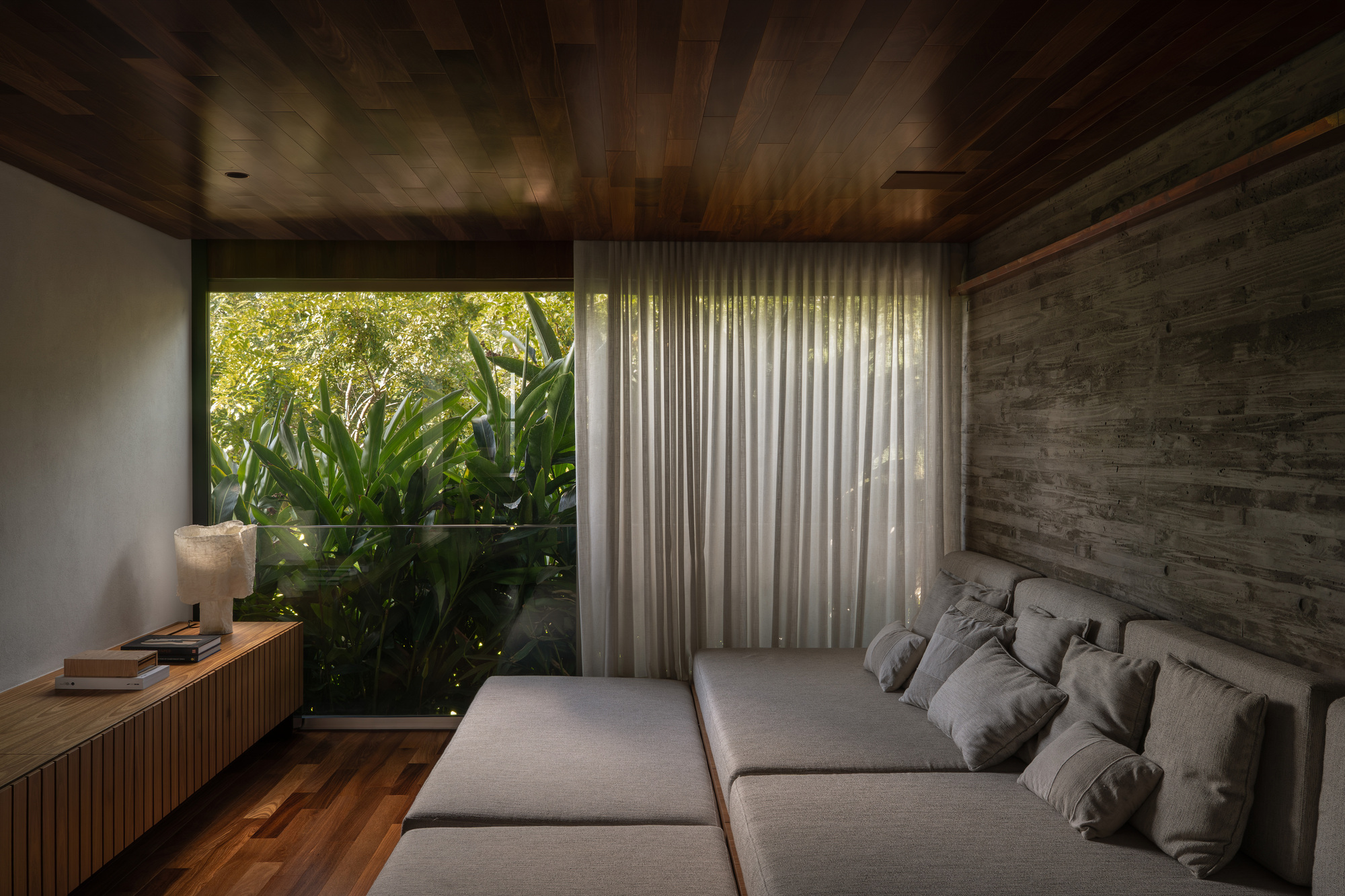 Lounge with a low grey sofa, sheer curtain and timber ceiling facing a glass-guarded view of tropical leaves