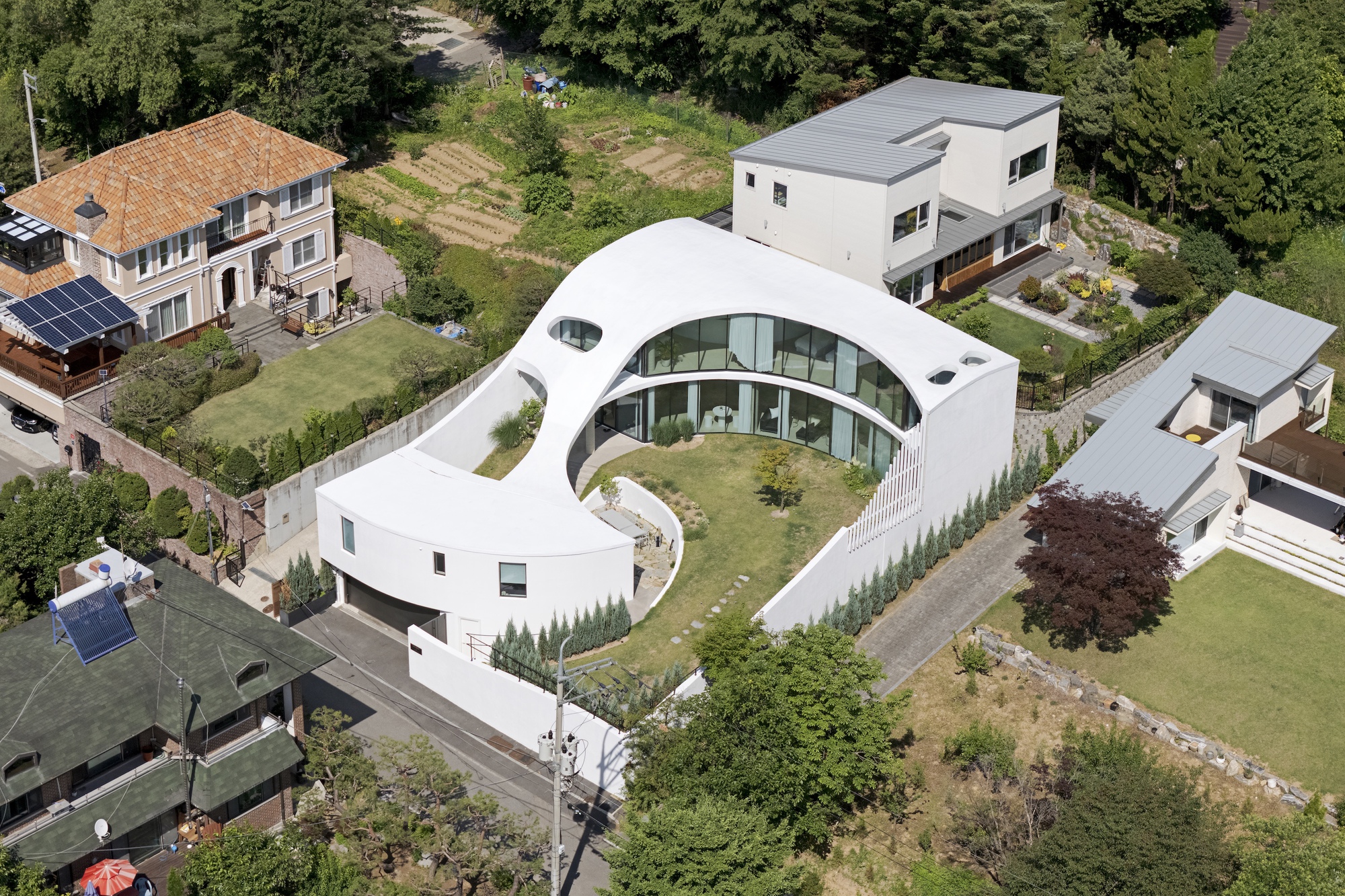 Aerial view of the white crescent-shaped house wrapping a lawn courtyard, surrounded by neighboring houses and trees on a sunny summer day
