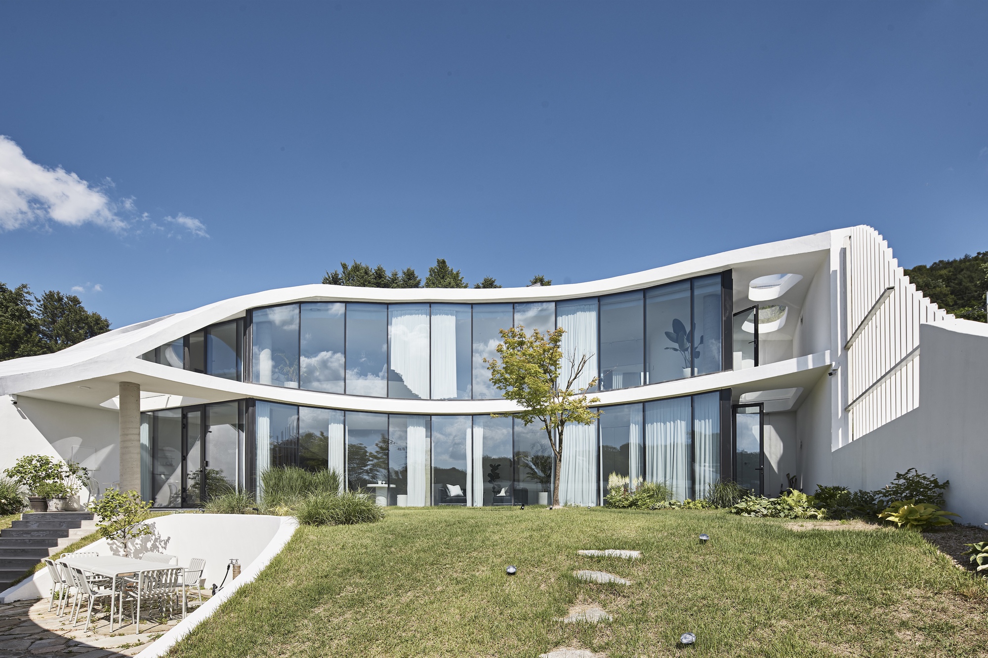 Curved glass facade under a sweeping white roof band, seen across a sloping lawn with stepping stones beneath a deep blue sky