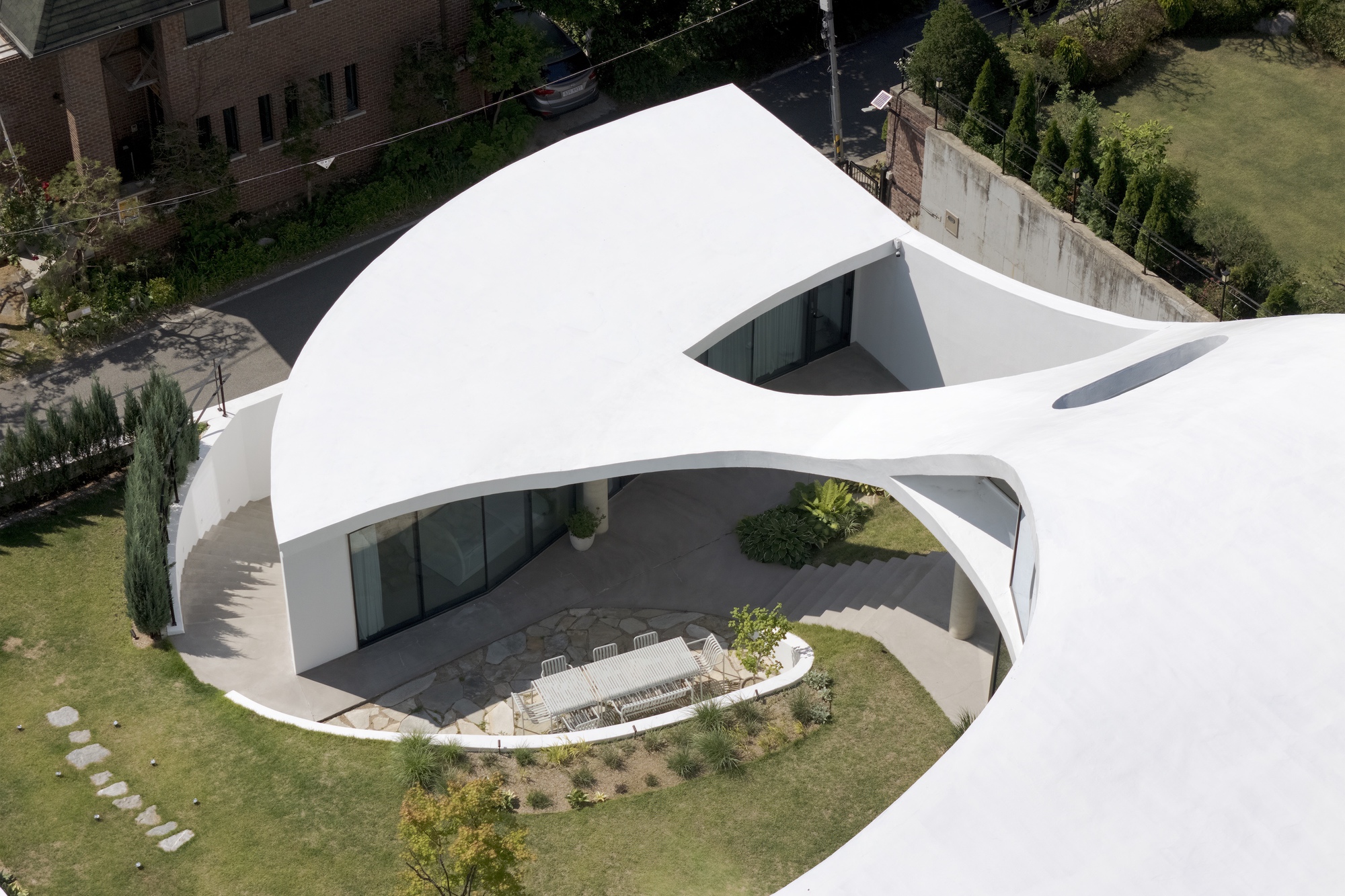 Aerial view of the lobed white roof sheltering a paved terrace with a dining table, beside a lawn and cypress hedge