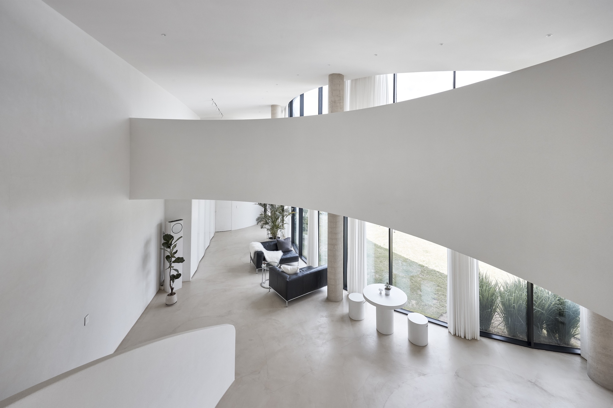 Upper-level view of a curved white balcony wall above a living area with concrete columns, sheer curtains and a grassy view outside