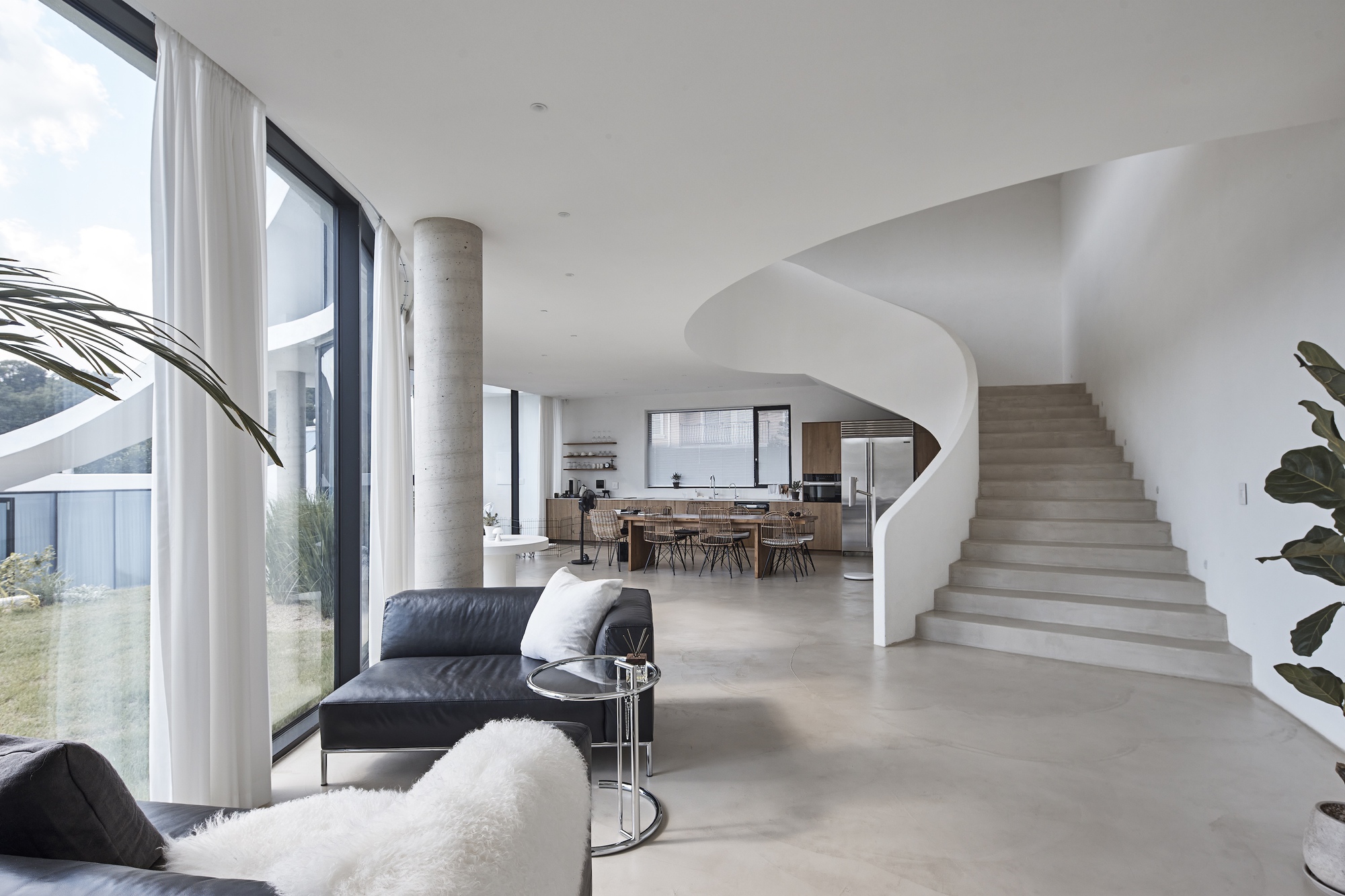 Open living space with a sculptural white spiral stair, concrete column and kitchen beyond, lit by daylight through full-height curtained glazing