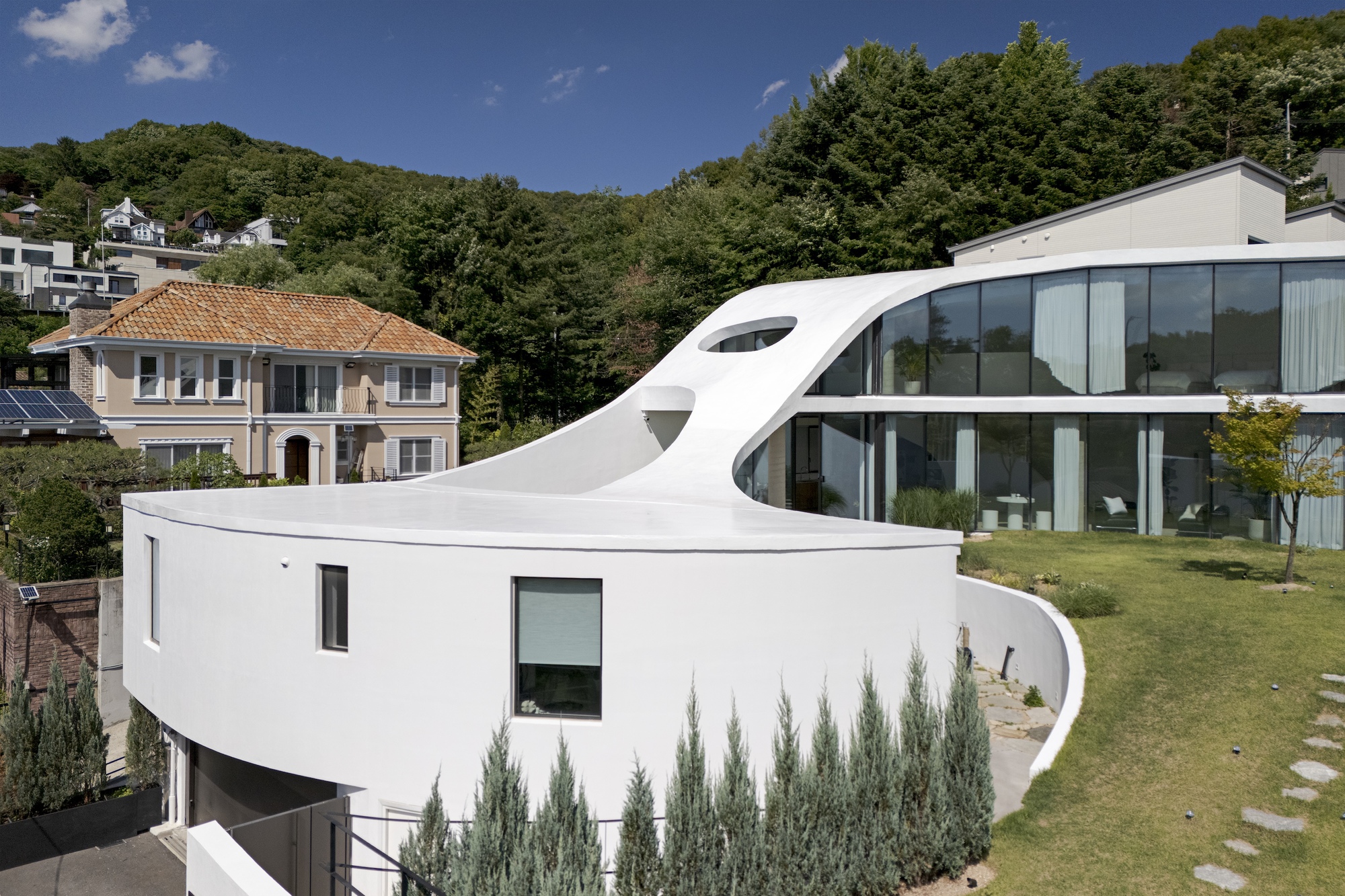 Rounded white volume and glazed upper floor above a lawn with stepping stones, with a wooded hillside and a tile-roofed house behind