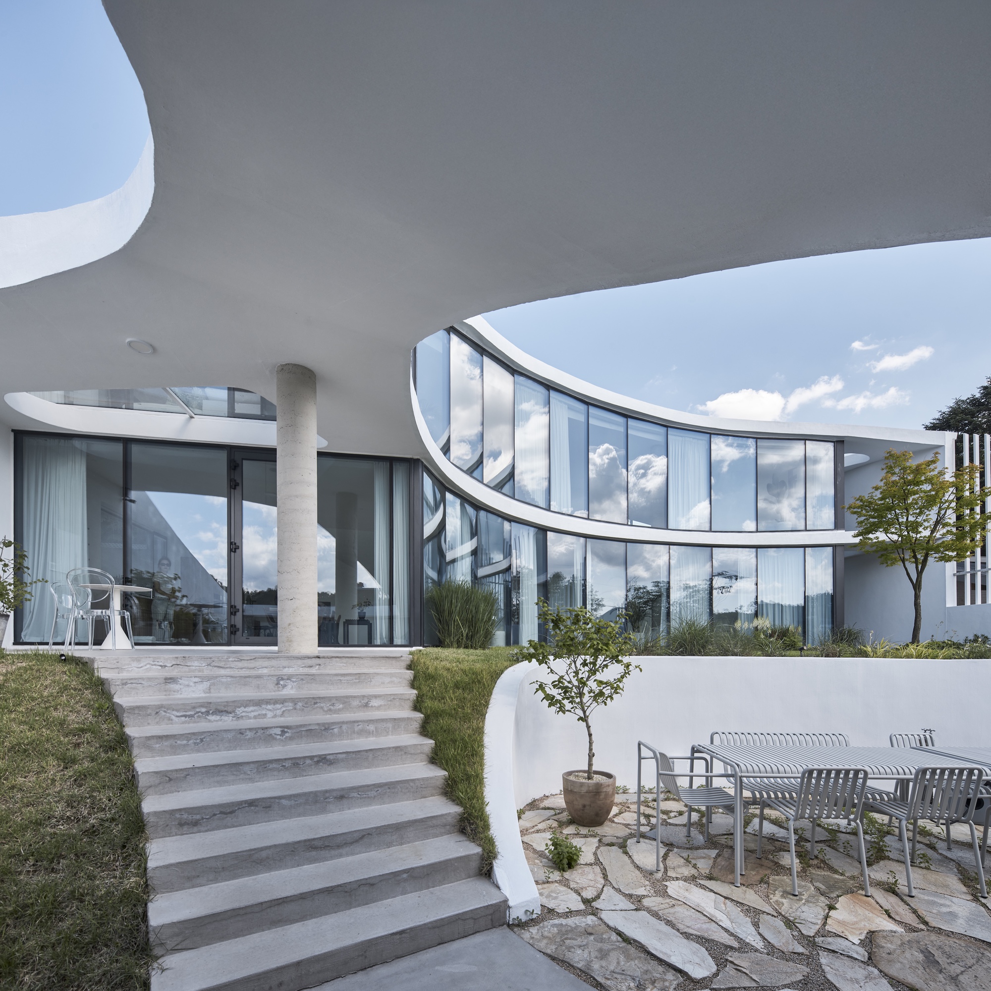 Concrete steps rising beside a round column and curved glass wall, with a patio table on stone paving under scattered clouds