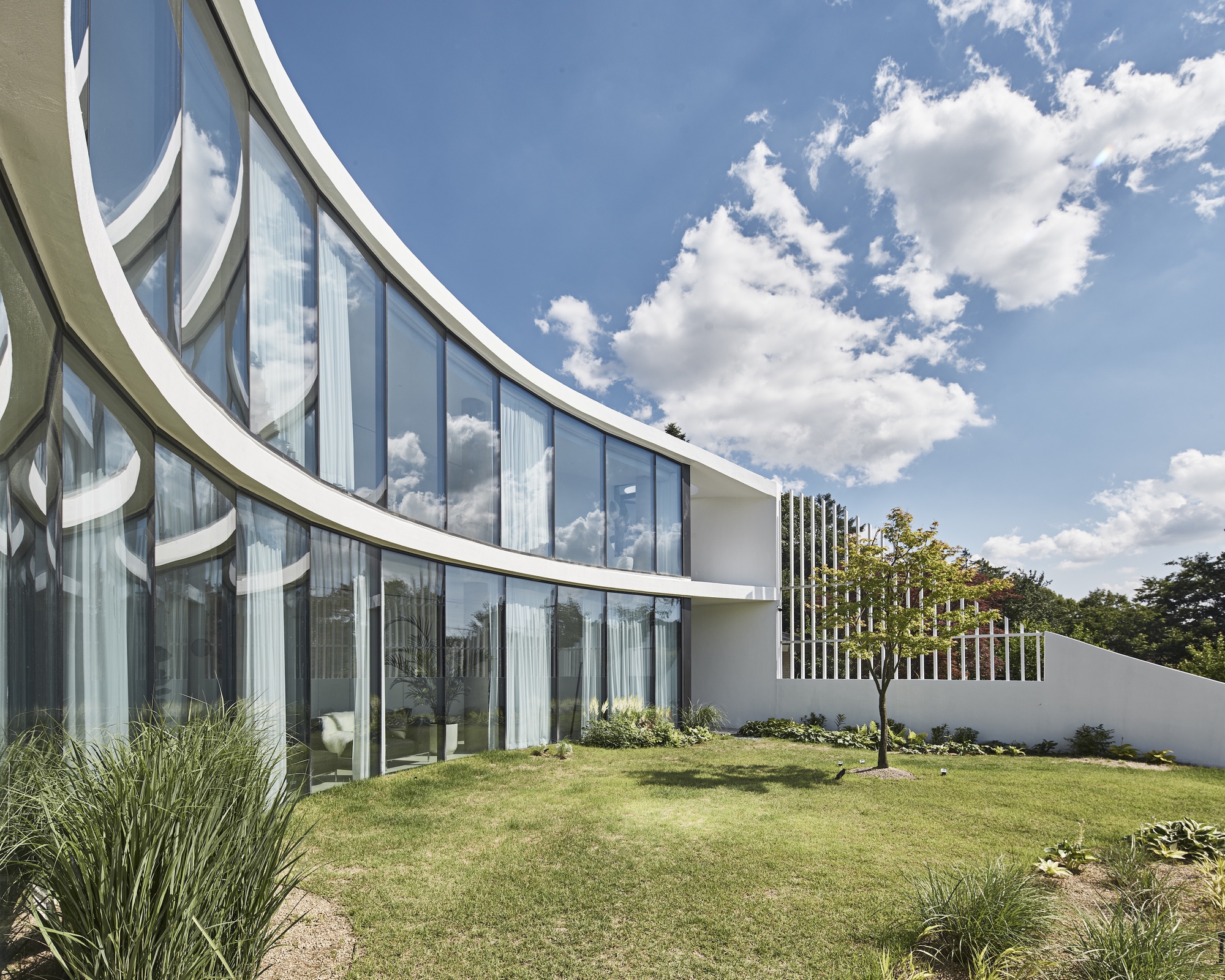 Curved glazed facade with white edge bands facing a lawn, a slatted screen and a young tree under a cloudy sky