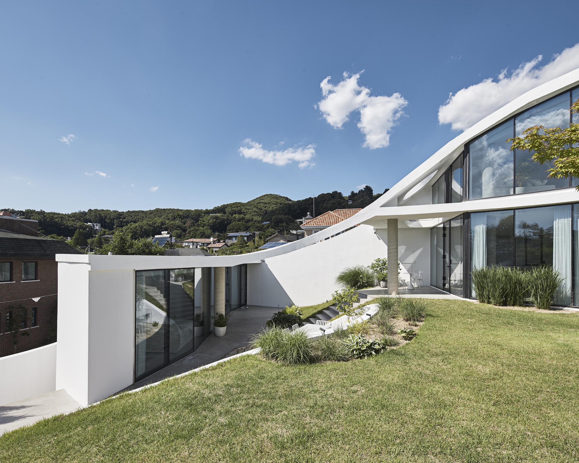 White sweeping roof edge over a glass wall and a sloped lawn, with wooded hills beyond on a clear day