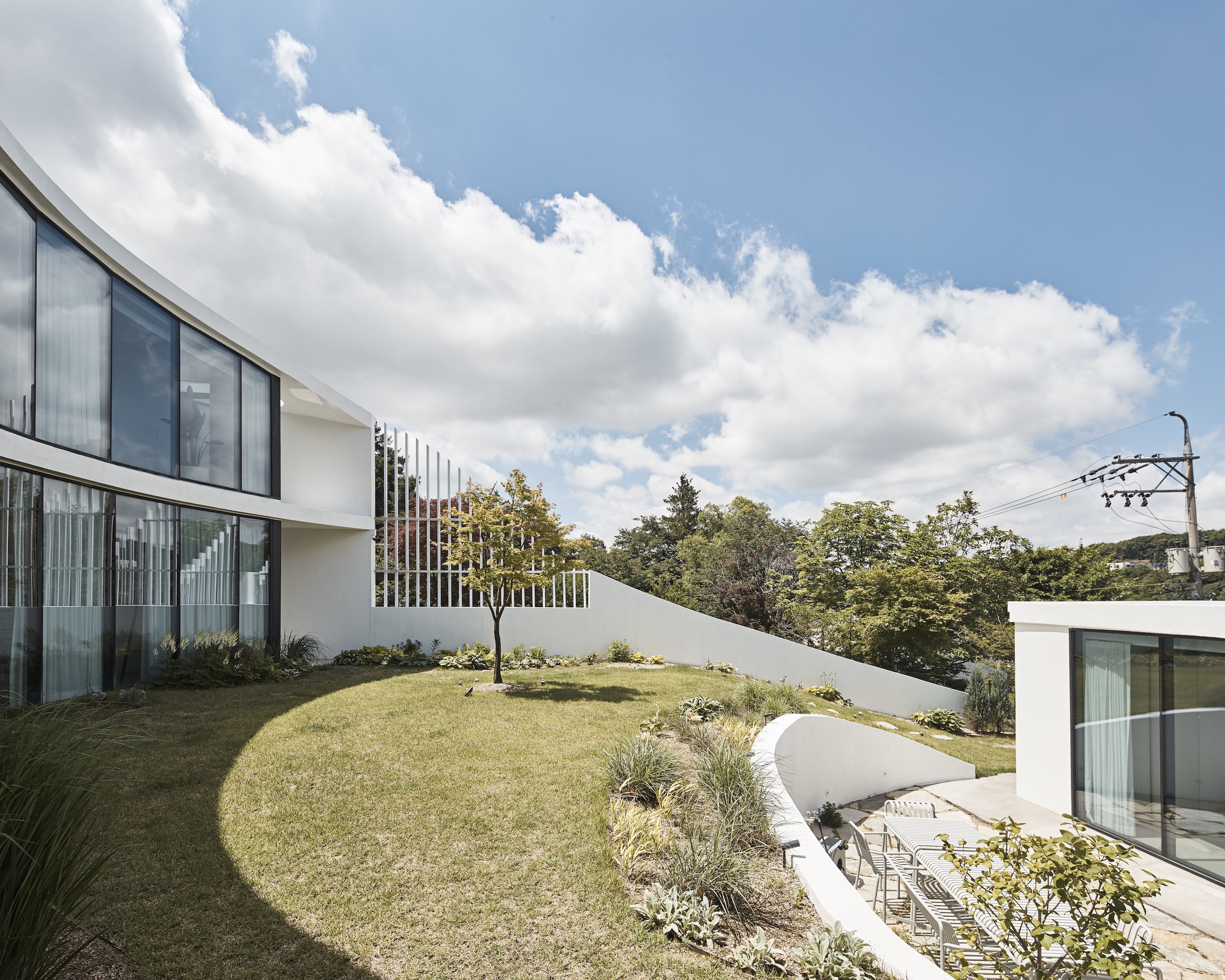 Curved glass wing and white retaining wall around a sunlit lawn, with a small tree and a sunken terrace with chairs