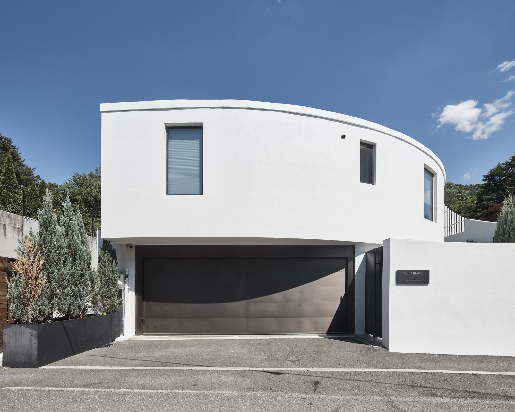 Street facade of a curved white upper volume above a dark garage door, with a planter of conifers in sunlight