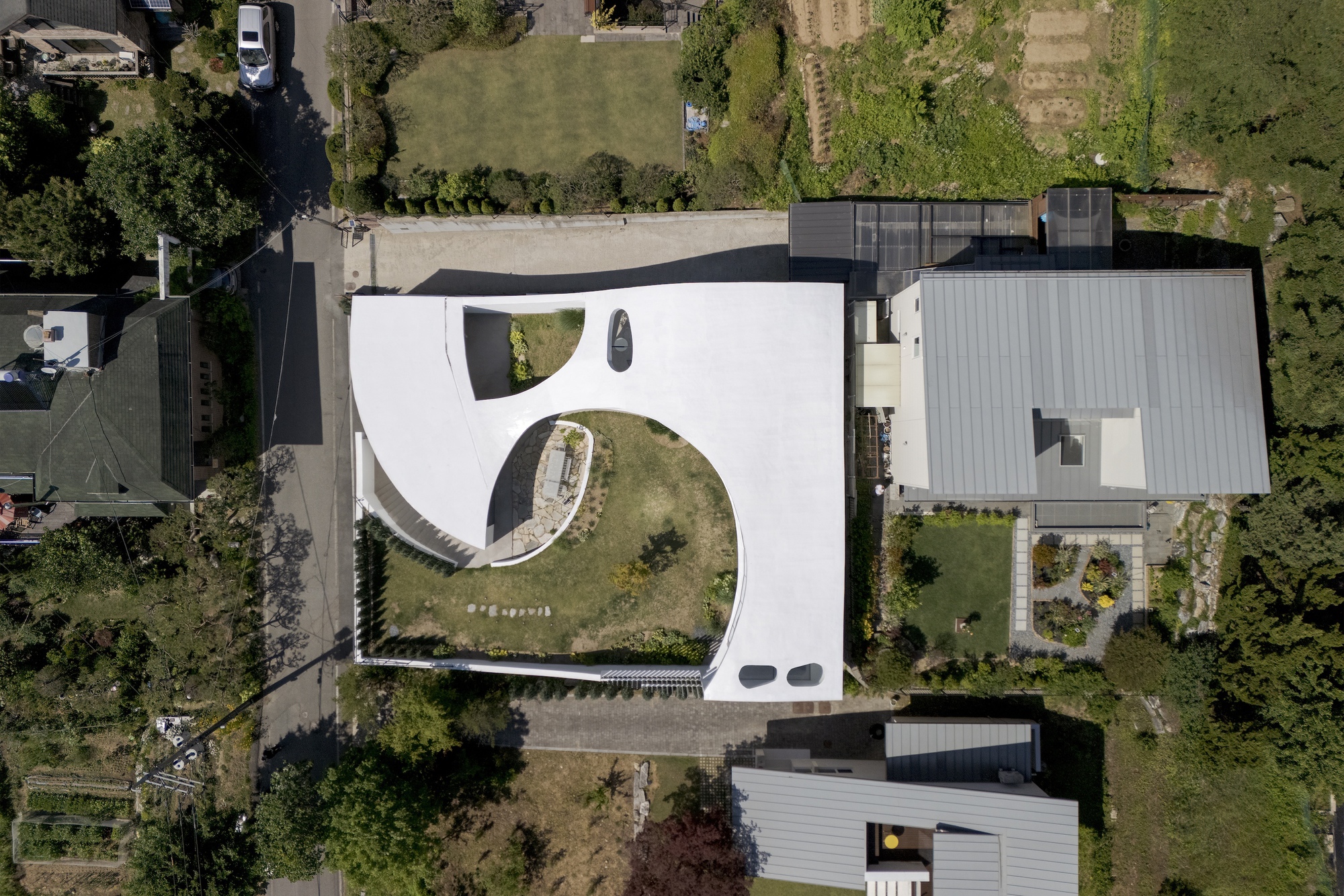 Top-down view of the white curved roof wrapping a grassy courtyard, beside neighboring metal-roofed houses in bright daylight