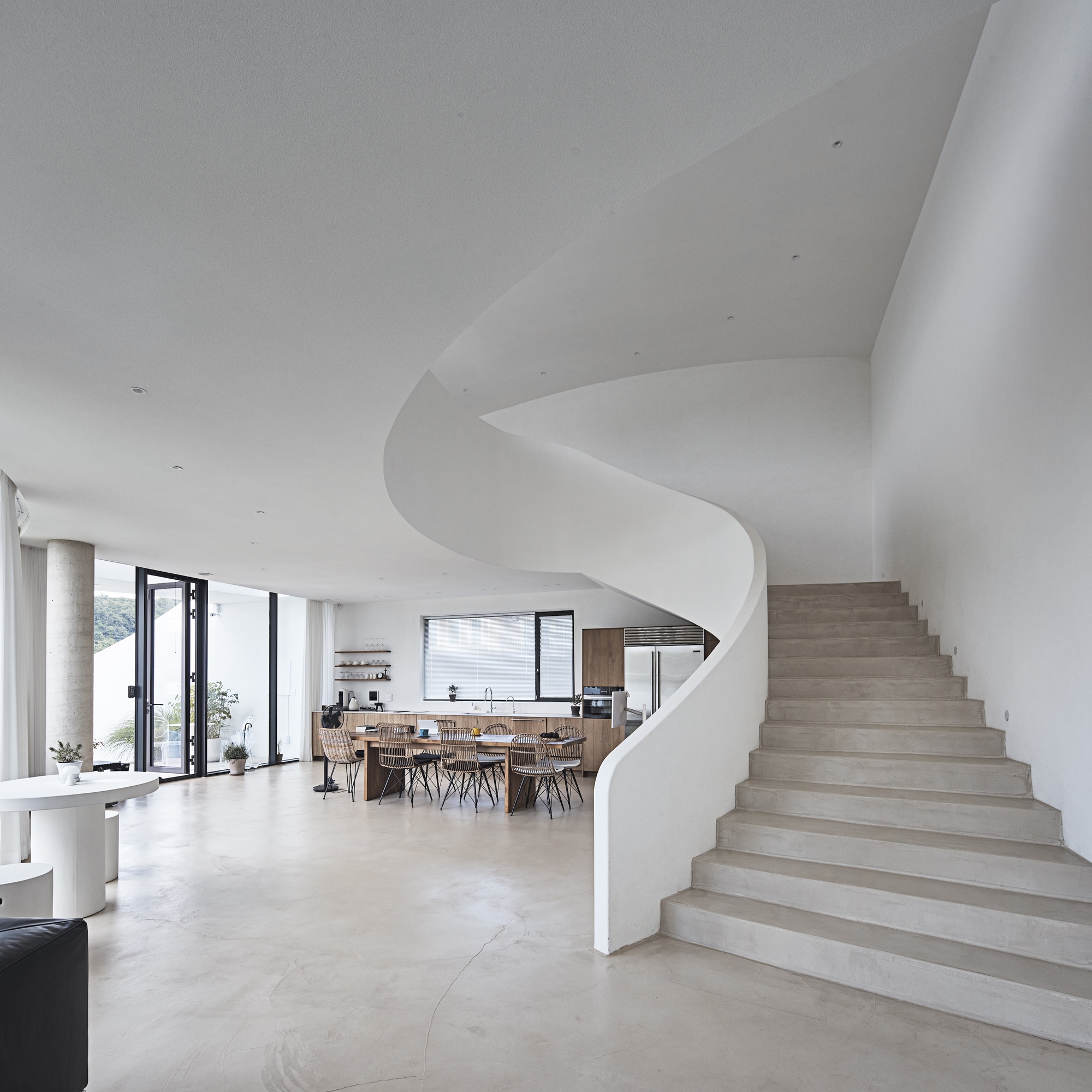 Sweeping white spiral stair beside an open kitchen and dining table, with a round concrete column near the glazed wall