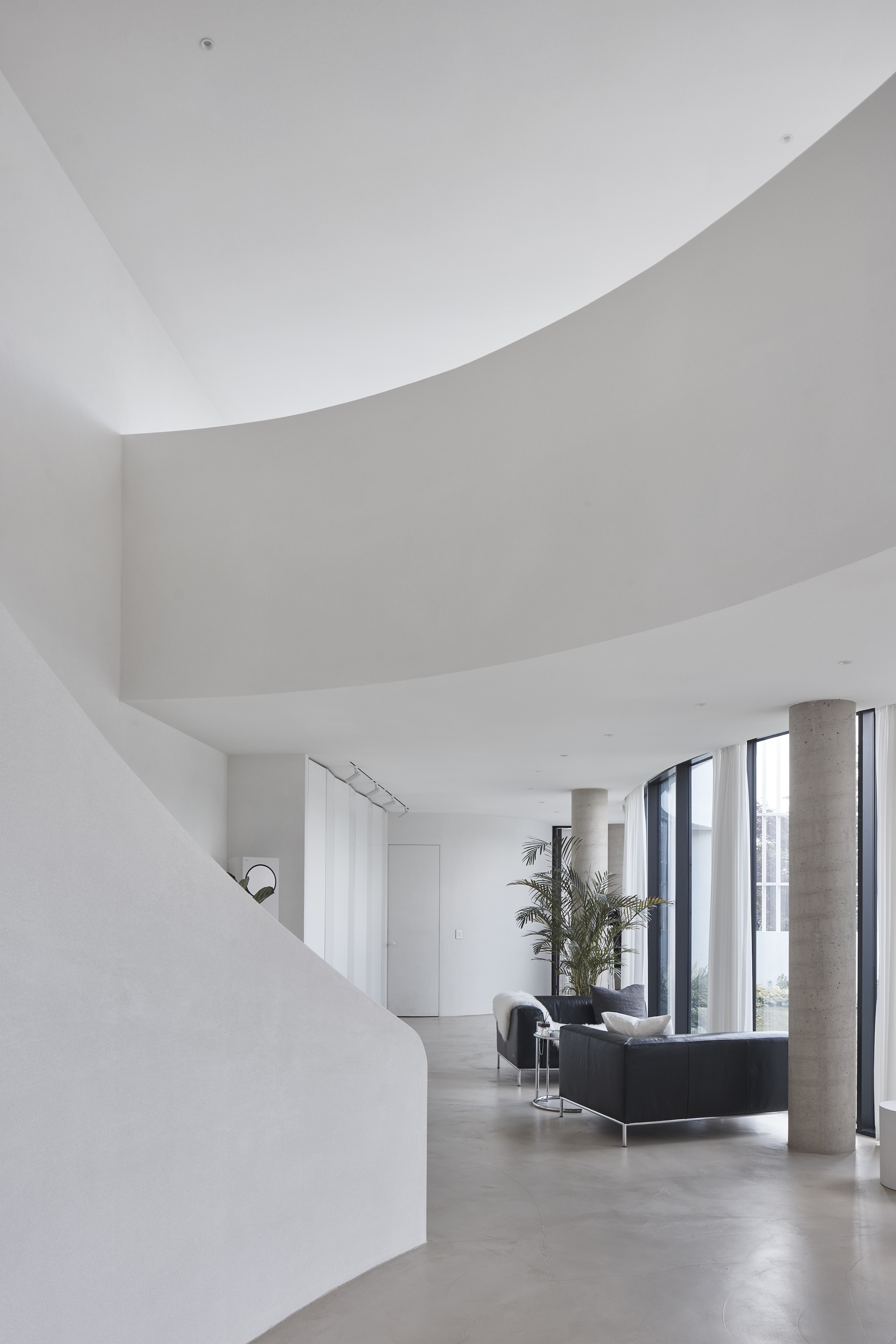 Curved white balcony edge above a lounge area with black sofas, a palm and concrete columns by the glass