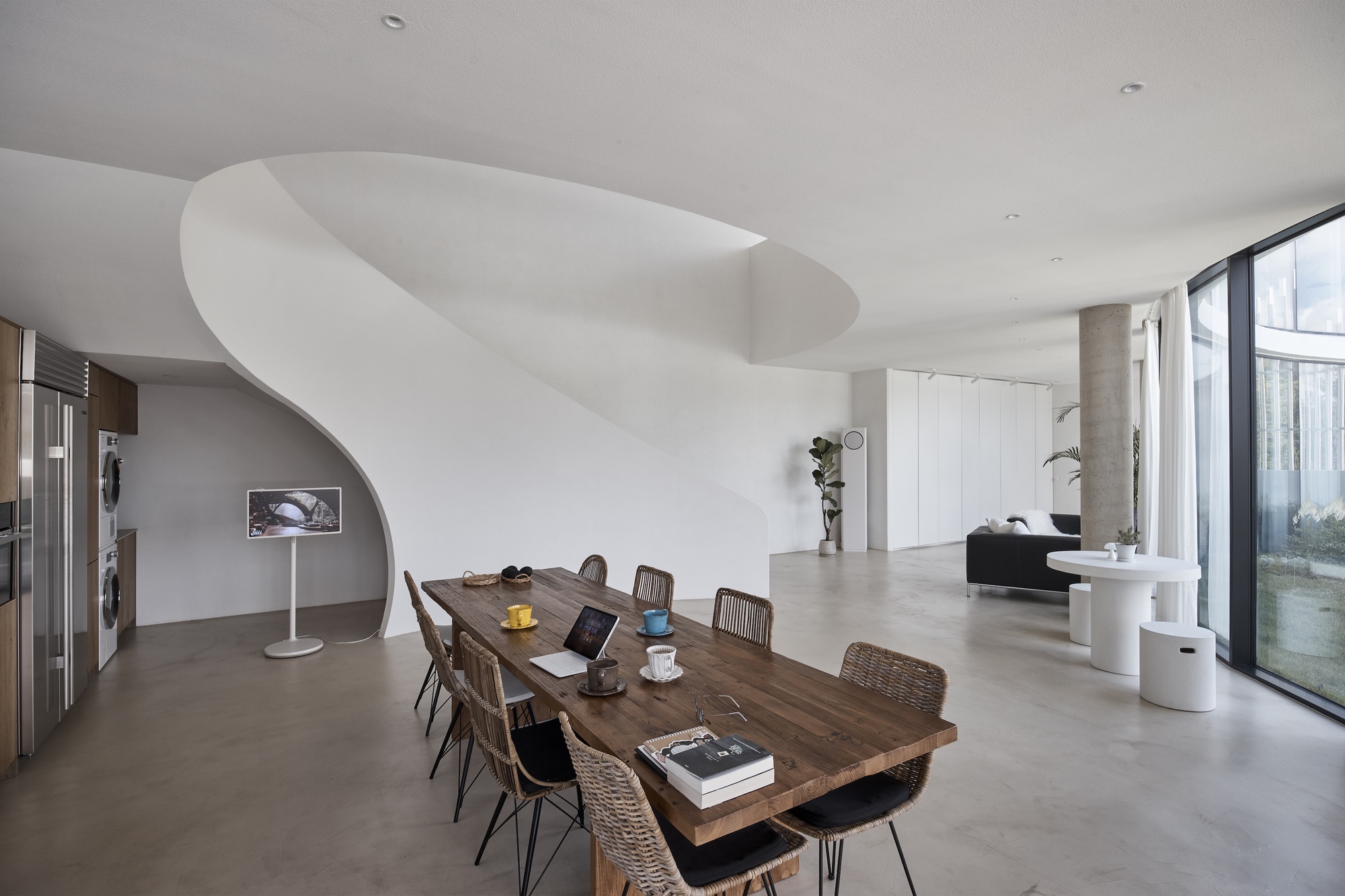 Long wooden dining table in front of the spiral stair, with cups, a laptop and a curved glass wall