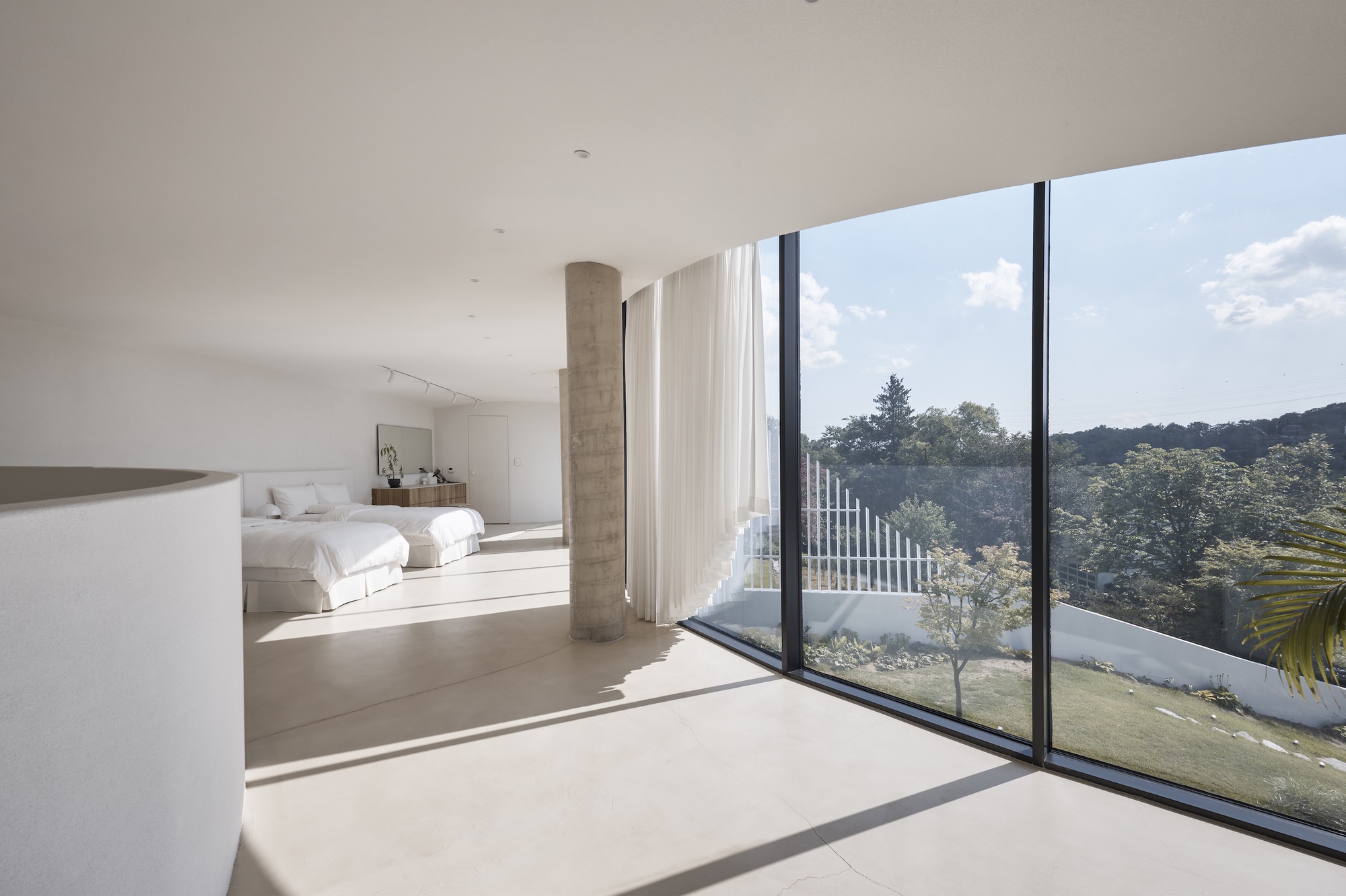 Bedroom with a concrete column and white curtain beside a curved glass wall, sunlight falling across the floor and hills beyond