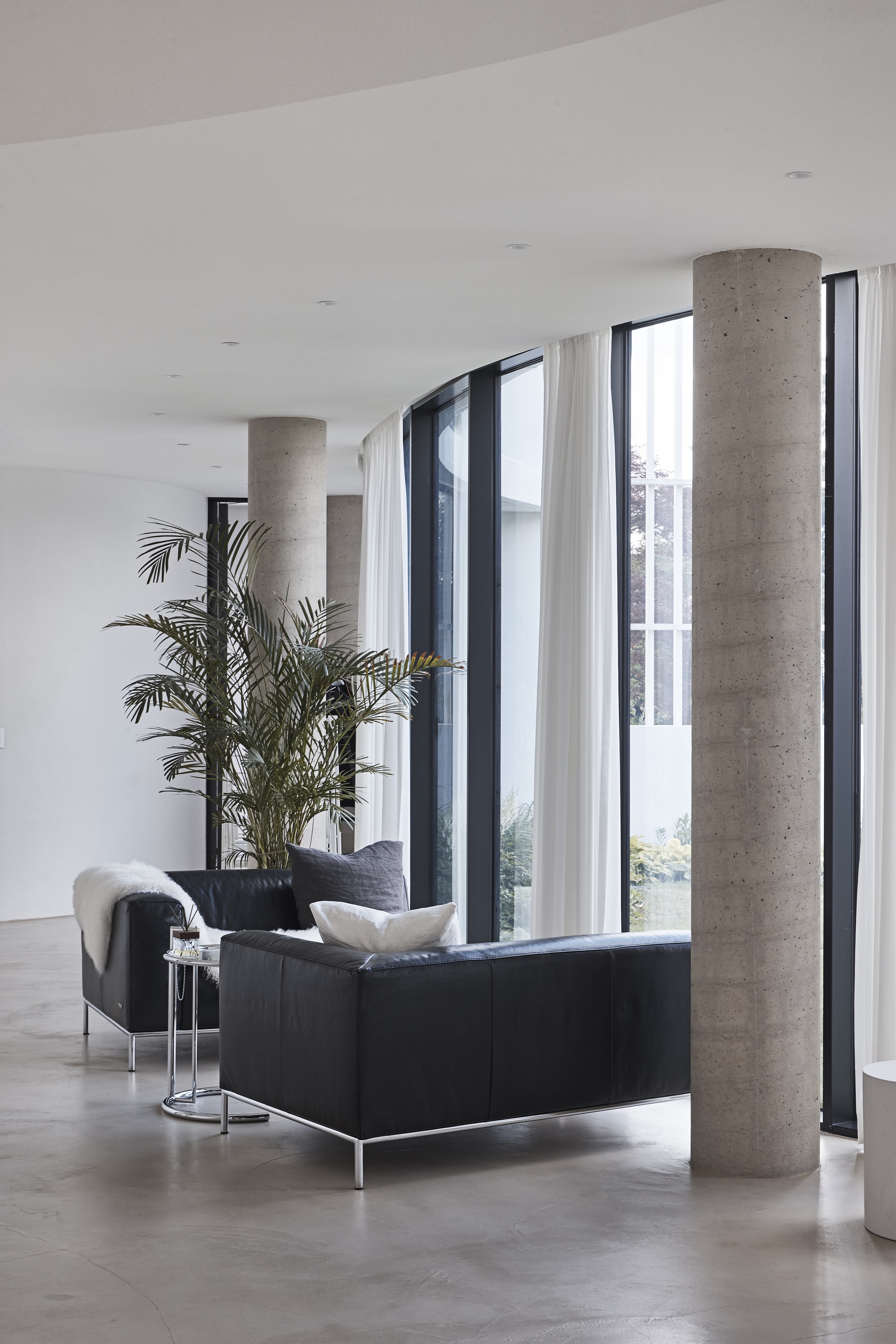 Board-formed concrete column beside a black leather sofa and potted palm, with sheer curtains along the curved glazing