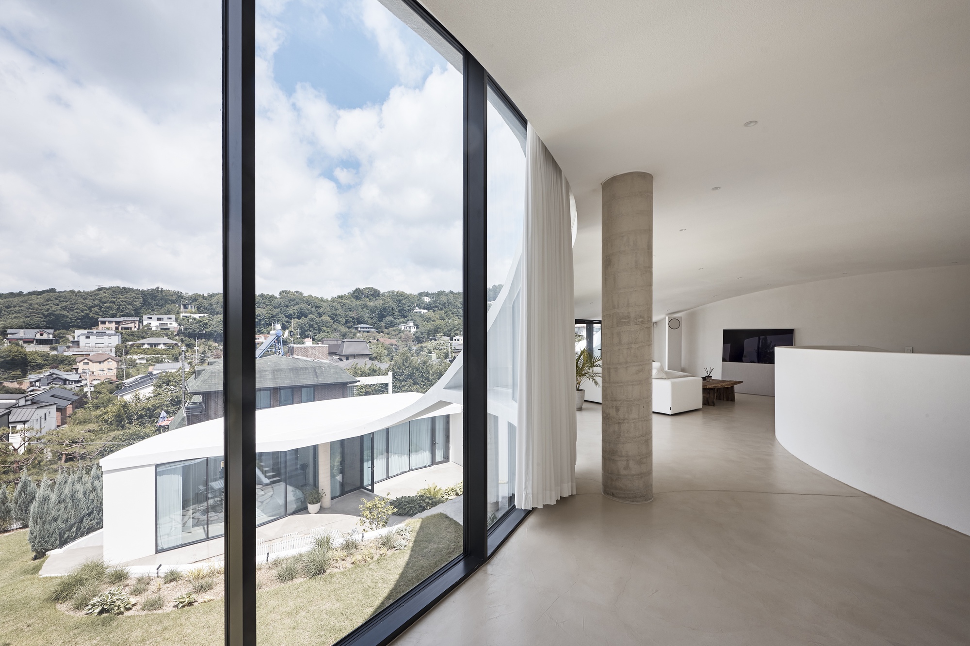 View through the curved glass wall past a concrete column to a sloping lawn and hillside houses under cloud