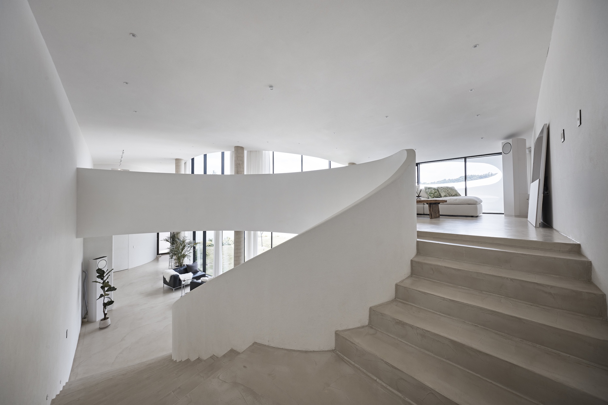 Curved white plaster stair with wide concrete treads rising toward a lounge, with a potted fig by the lower landing