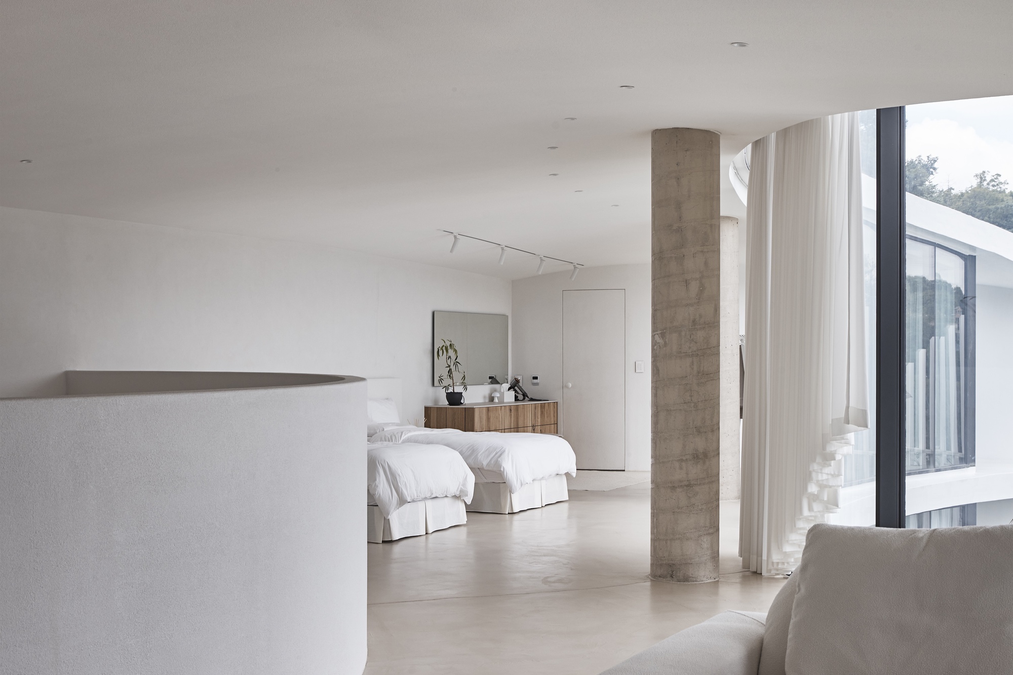 Open sleeping area with twin white beds beside a concrete column, a curved parapet wall in the foreground and daylight from the glazing