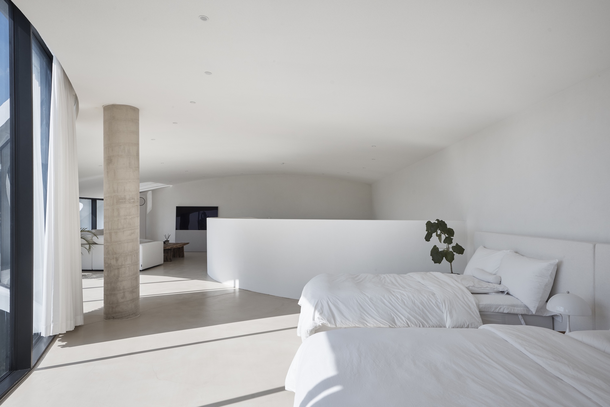 Bedroom with white linen beds under a vaulted ceiling, sunlight striping the floor beside a concrete column and sheer curtain